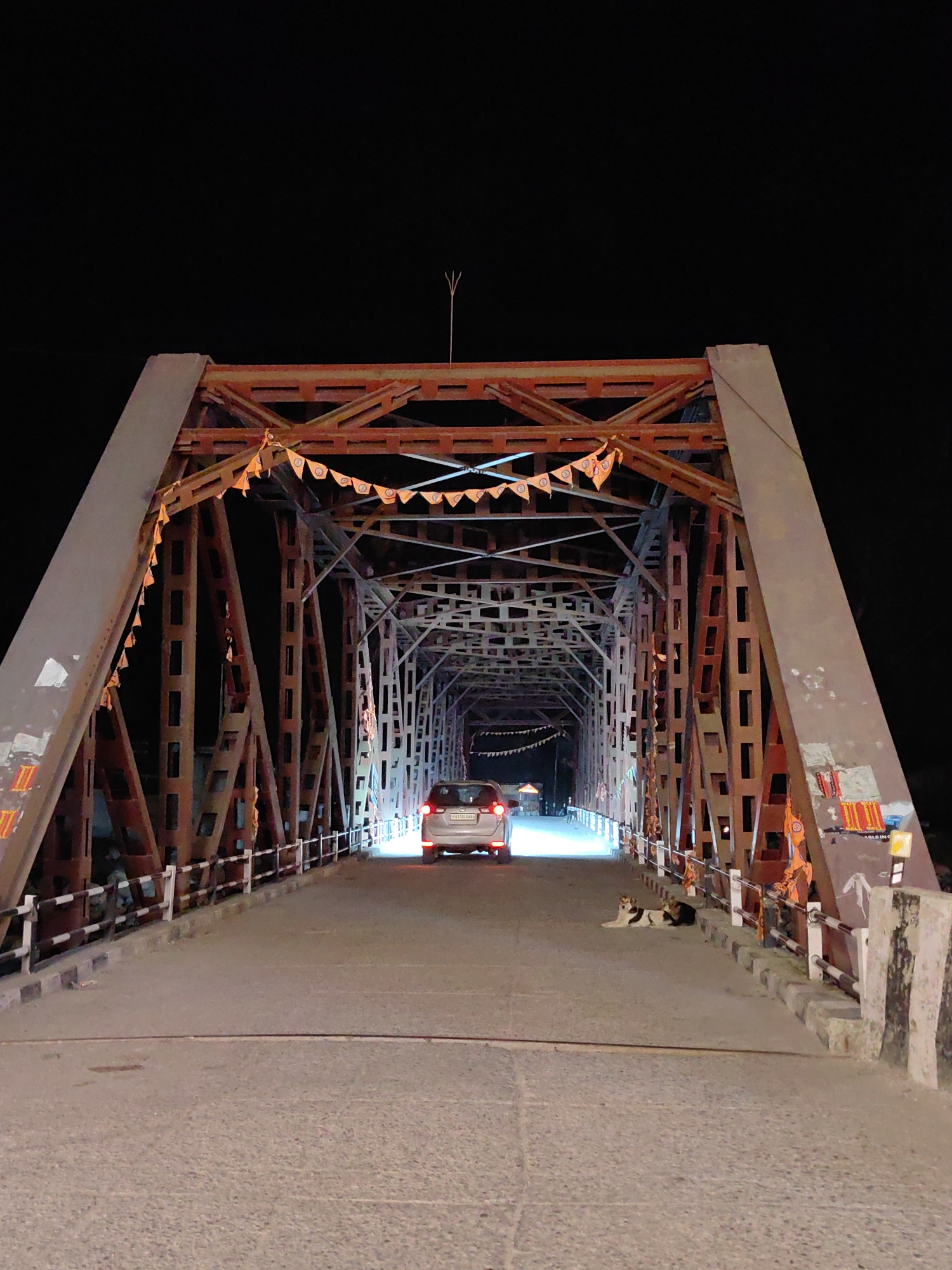 This image depicts a nighttime scene of a metal truss bridge in Kulang, India. The bridge is illuminated by artificial lights, casting a warm glow on the structure and the road. A silver car is visible driving away from the camera towards the end of the bridge. To the right of the car, two dogs are resting on the road. The bridge is adorned with small orange flags strung across its upper section, suggesting a local celebration or religious observance. The overall environment is dark, with the night sky visible above the bridge's structure.