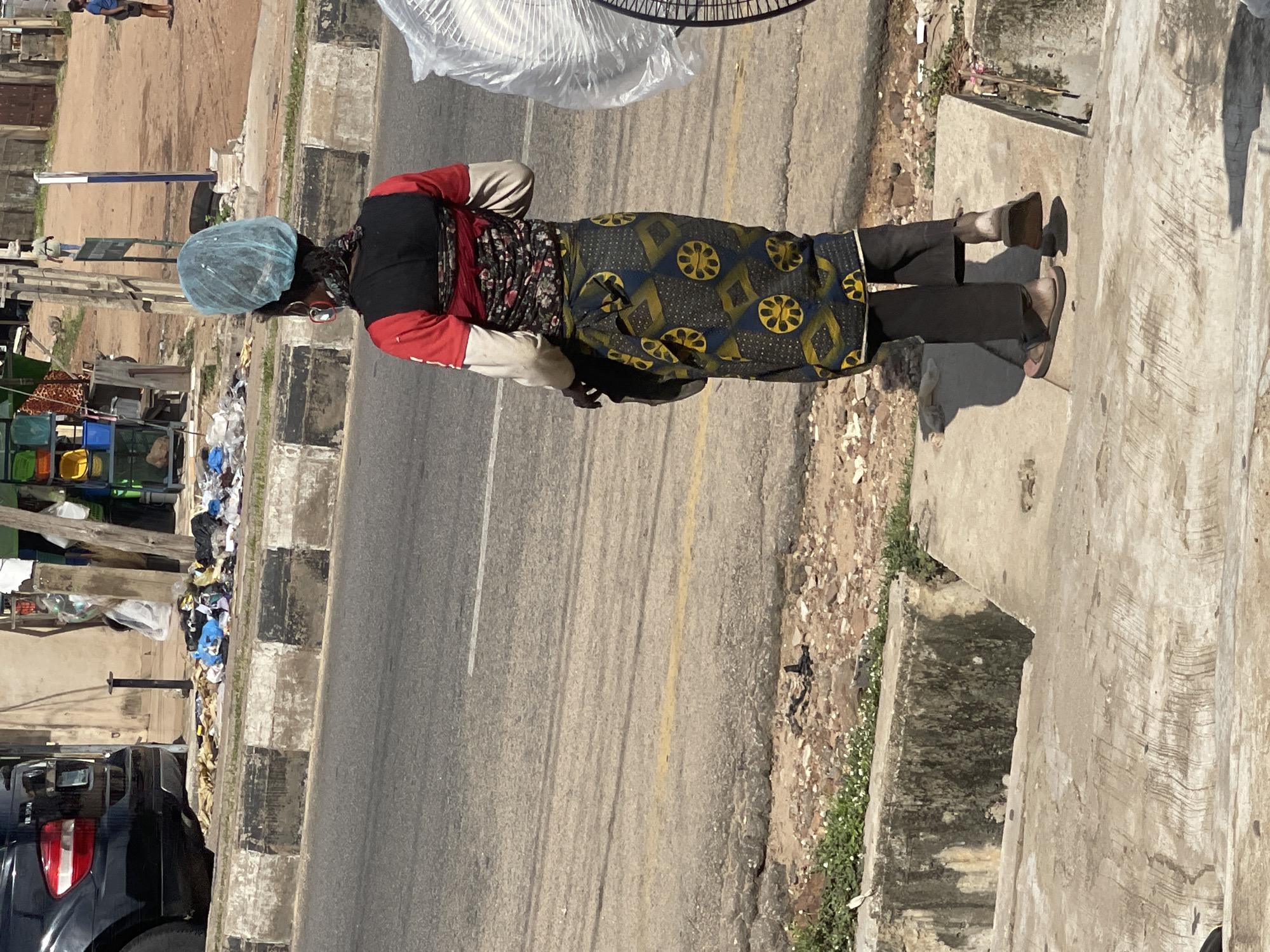 A full-length shot shows a person standing on a paved sidewalk next to a street. The person is wearing a blue hairnet, a red long-sleeved shirt with a black and floral print section, and black pants. They are also wearing dark shoes and sandals. The street is paved and has a yellow line and a white line marking lanes. The sidewalk is made of concrete and has some debris on it, including trash. Behind the person, there is a cluttered area with various items, including what appear to be market stalls, baskets of goods, and more trash. A black vehicle is parked on the left side of the image, near the sidewalk. The scene appears to be outdoors during the daytime, with bright sunlight casting shadows. The overall impression is of a street scene in a busy urban or semi-urban area, possibly a market or roadside vendor location, with a focus on the person and their surroundings. There is no visible text in the image.