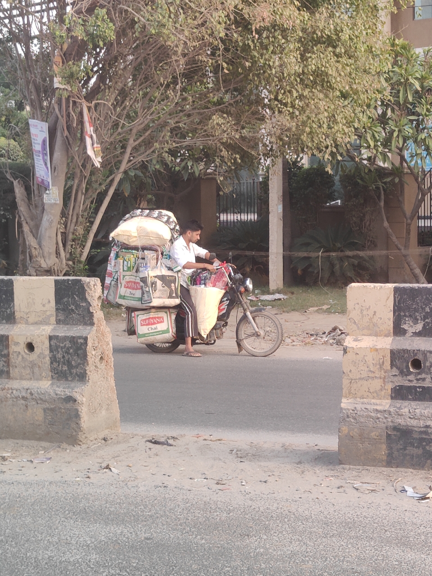 A man rides a motorcycle laden with goods through a street in Gurgaon, India. The motorcycle is piled high with bags of tea and other items. The man is wearing a white shirt and dark pants. He is looking forward as he rides. The street is lined with trees and buildings. There is a concrete barrier on the left side of the road. The sky is overcast.