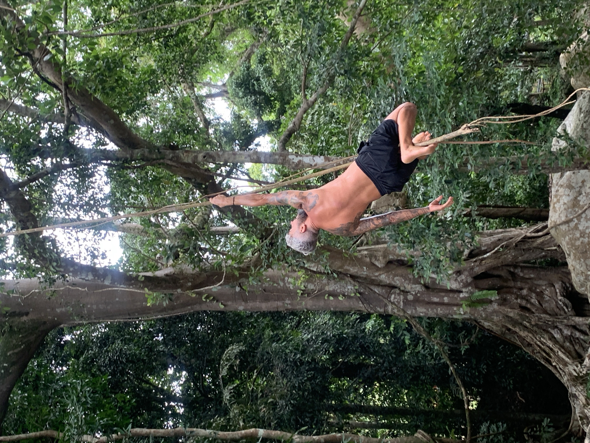 In the Ko Samui District of Thailand, a man with tattoos on his arms and back is captured mid-air, swinging from a thick vine in a lush, green jungle. He is wearing black shorts and is balancing himself using his feet on the vine. The background is filled with dense foliage and large trees, with sunlight filtering through the canopy. The scene appears to be a natural outdoor environment.