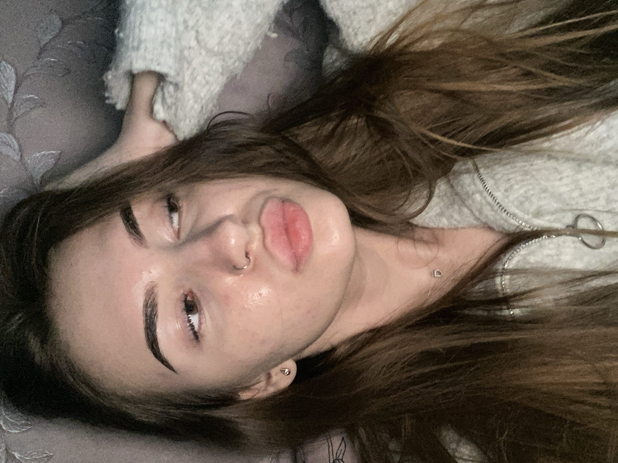 A close-up, top-down shot captures a young woman lying down indoors. Her head is tilted, with her face oriented towards the upper left of the frame. She has long, dark brown hair spread around her head and shoulders, blending with a light-colored, textured sweater or hoodie that has a zipper detail visible on the right. Her skin appears dewy, and her dark eyebrows are well-defined. She is looking slightly upward and to the left with a somewhat heavy-lidded gaze, her lips slightly pursed in a pout.
Notable features include a small silver hoop piercing through her septum, a small gold-toned stud earring in her left earlobe, and a delicate silver necklace with a tiny pendant resting on her collarbone. The background consists of a dark, patterned fabric or bedding on the left, possibly a pillow or headboard, and the light-colored, fuzzy material of her clothing on the right. The lighting is soft and ambient, suggesting an indoor setting. There is no visible text.