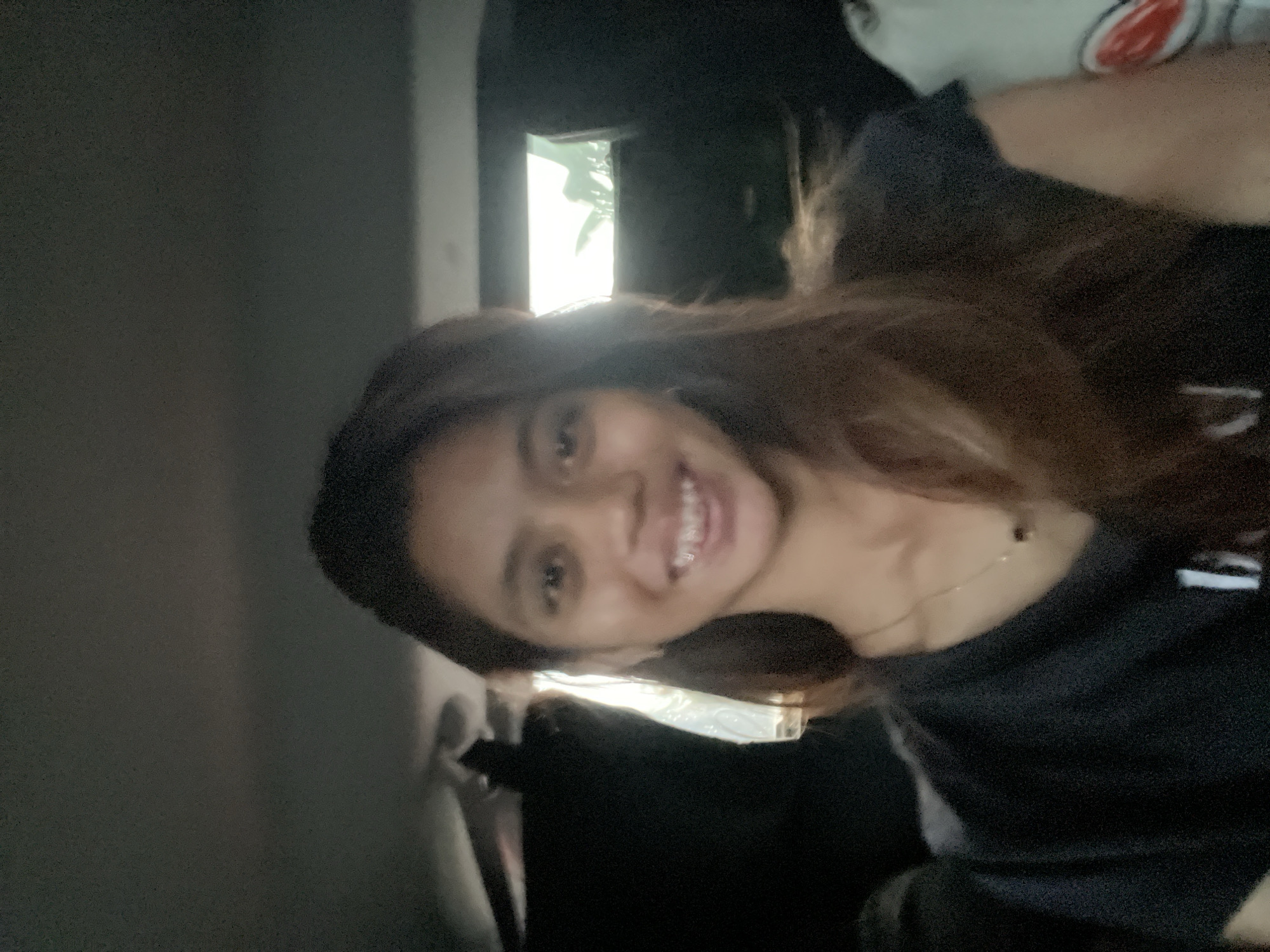 A young woman is smiling at the camera in an interior space. She has medium-length brown hair and is wearing a dark-colored shirt and a thin necklace. The setting appears to be a vehicle, as there is a car door frame visible. Bright light is coming in from a window behind her. The location is Davao City, Philippines.