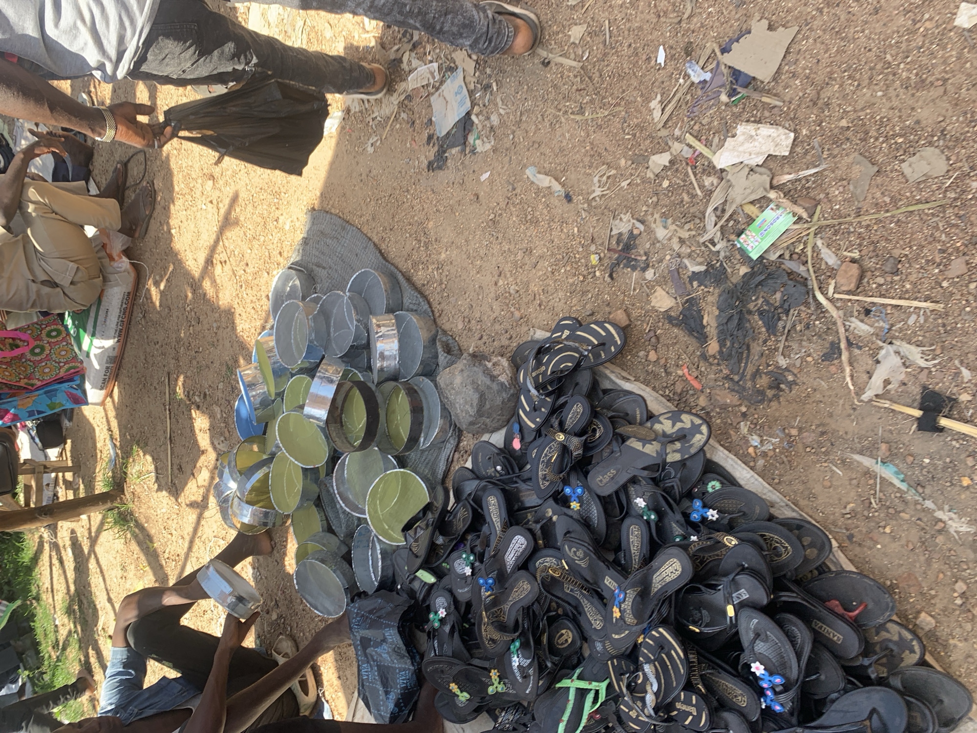 In Boi A, Nigeria, a scene unfolds at what appears to be an outdoor market or roadside stall. The sun casts long shadows, suggesting it is either early morning or late afternoon. Several individuals are present, their legs and arms visible as they engage with merchandise. One person, wearing dark pants and a light-colored shirt, is holding a black plastic bag. Another person, in light-colored pants, is seated on the ground, holding a cylindrical object.
The ground is dusty and covered with a mixture of dirt and debris, including bits of paper and plastic. A prominent display of items includes a large pile of black flip-flops, many adorned with decorative elements like flowers and gold accents. Some of the flip-flops have visible text, such as "Class" and "VAINVE." Alongside the footwear, there is a collection of metal rings, some with green interiors, stacked on a grey tarp.
Other objects in the scene include colorful patterned fabrics, a white bag with text that is partially legible, and various pieces of discarded material scattered across the ground. The overall impression is one of a bustling marketplace where goods are being sold or prepared for sale in an open-air environment. The mood seems neutral, focused on the transactional nature of the setting.
