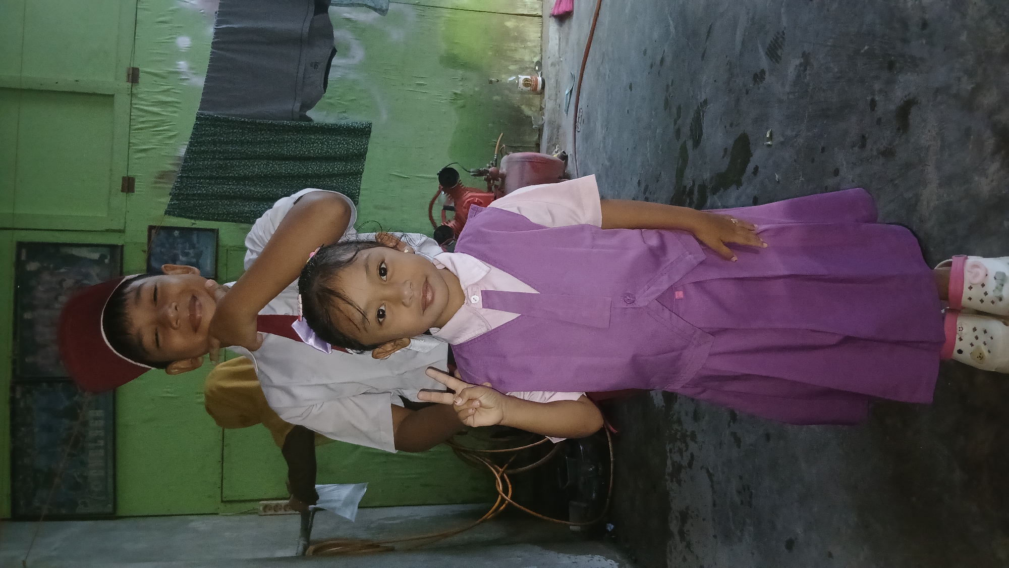 A young girl in a purple school uniform stands in the foreground, making a peace sign with her right hand. She is wearing a light pink collared shirt with a purple vest over it, a purple tie, and a pleated purple skirt. Her shoes are white with pink trim and decorated with cartoon characters. Behind her, a young boy wearing a red cap, white shirt, and red tie stands, smiling and resting his chin on his hand. He appears to be a student. In the background, there is a green wall with framed pictures, a clothesline with hanging garments, and some industrial equipment. The scene appears to be an outdoor or semi-outdoor area, possibly a schoolyard or a backyard, with a concrete floor. The overall impression is of children in school uniforms, possibly at a school event or on their way to school, in Keudejungkagajah, Indonesia.