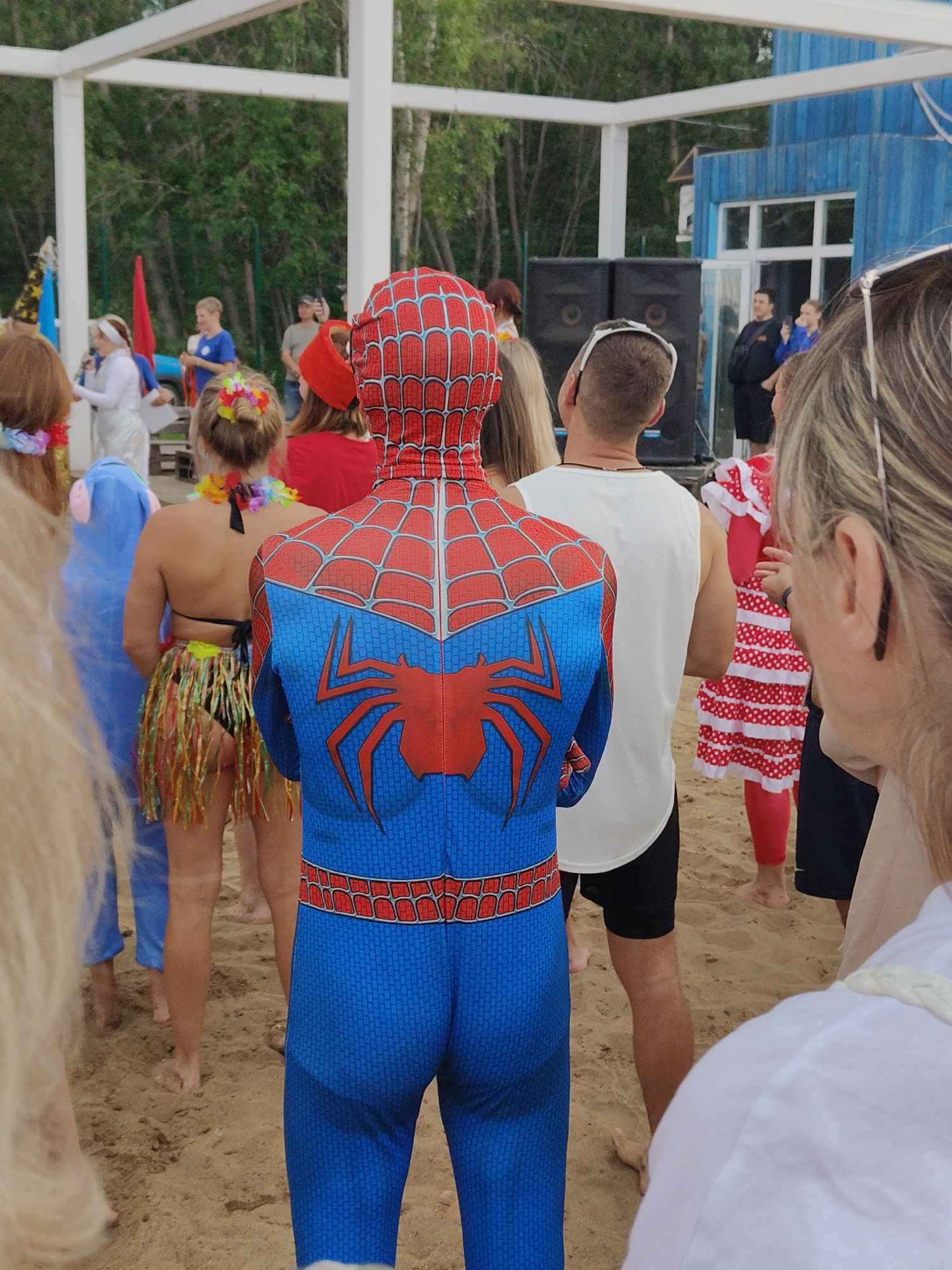 The image depicts a group of people on a sandy beach, with a wooden structure in the background and a blue building. In the foreground, a person is wearing a full Spiderman costume. To the left, another individual is dressed in a blue Eeyore costume. Behind them, a woman wears a bikini and a grass skirt. Several other people are visible in the background, some appearing to be dressed in festive attire, possibly for a party or event. Two large speakers are placed near the blue building. The overall atmosphere suggests a casual outdoor gathering.