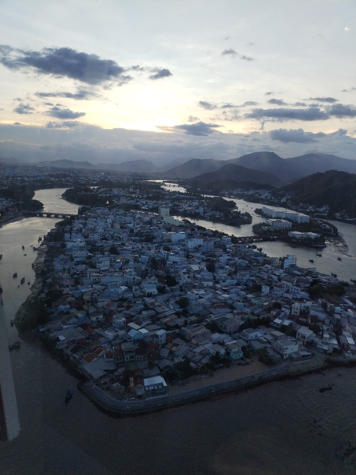 The image presents an aerial view of a dense residential area within Bac Nha Trang Ward, Vietnam. The area is characterized by tightly packed buildings with diverse roof colors, primarily composed of single-family homes. A significant river runs through the landscape, branching around islands. In the distance, mountains are visible under a sky with scattered clouds and a visible sunset. Boats are scattered along the river. The lighting suggests it is either early morning or late evening.