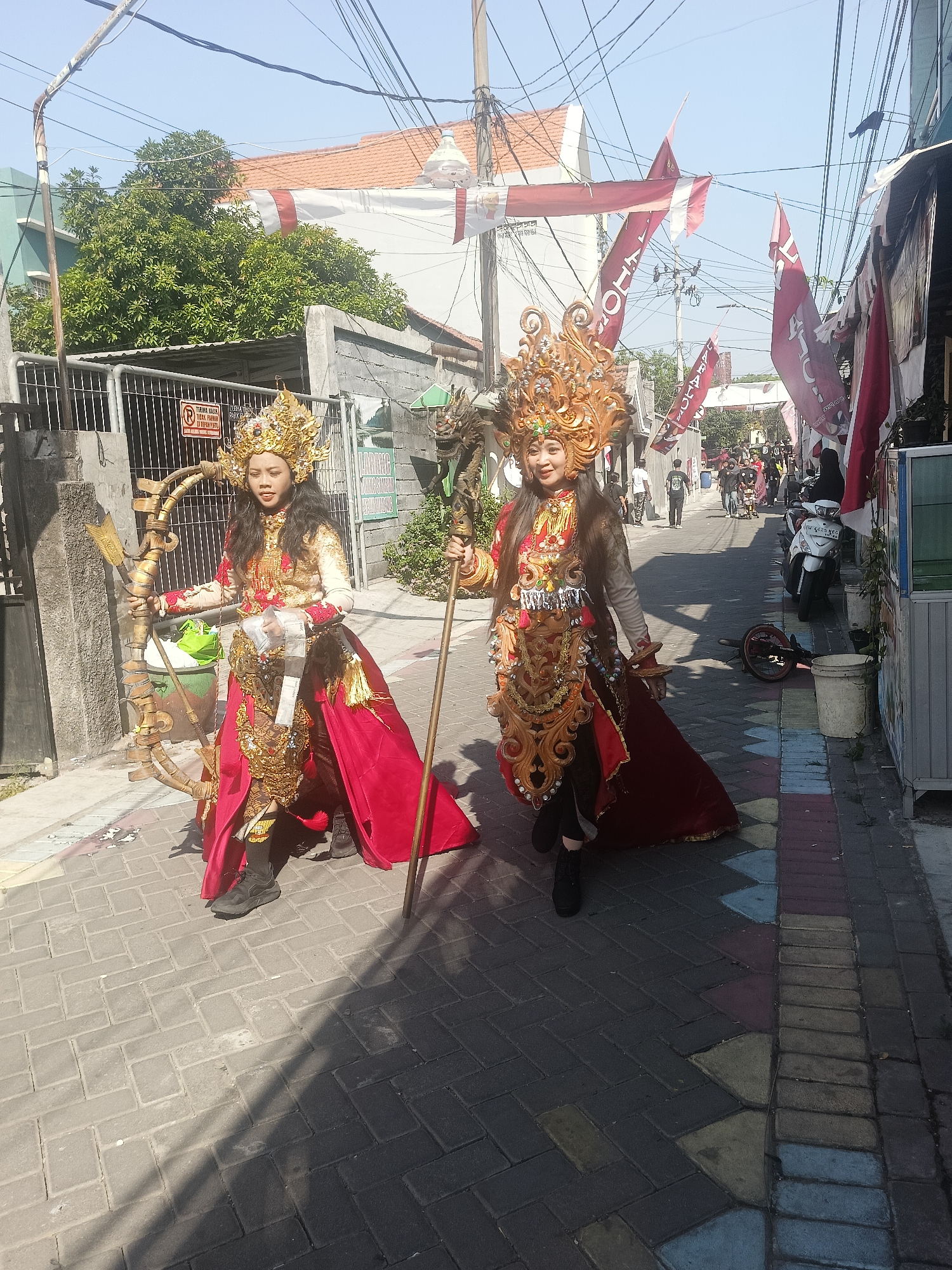 Two women, adorned in elaborate, traditional Indonesian costumes featuring golden headdresses, ornate gold and red garments, and flowing red capes, walk along a cobblestone street in Gedangan, Indonesia. The woman on the left holds a large, golden bow and arrow prop, while the woman on the right carries a golden staff. They are walking in the same direction on a street lined with buildings and overhead electrical wires. Red and white Indonesian flags and banners are visible, some hanging across the street and others attached to buildings, suggesting a celebration or holiday. In the background, other people are walking, and a motorcycle is parked on the side of the street. The scene is illuminated by bright sunlight, casting shadows on the ground.