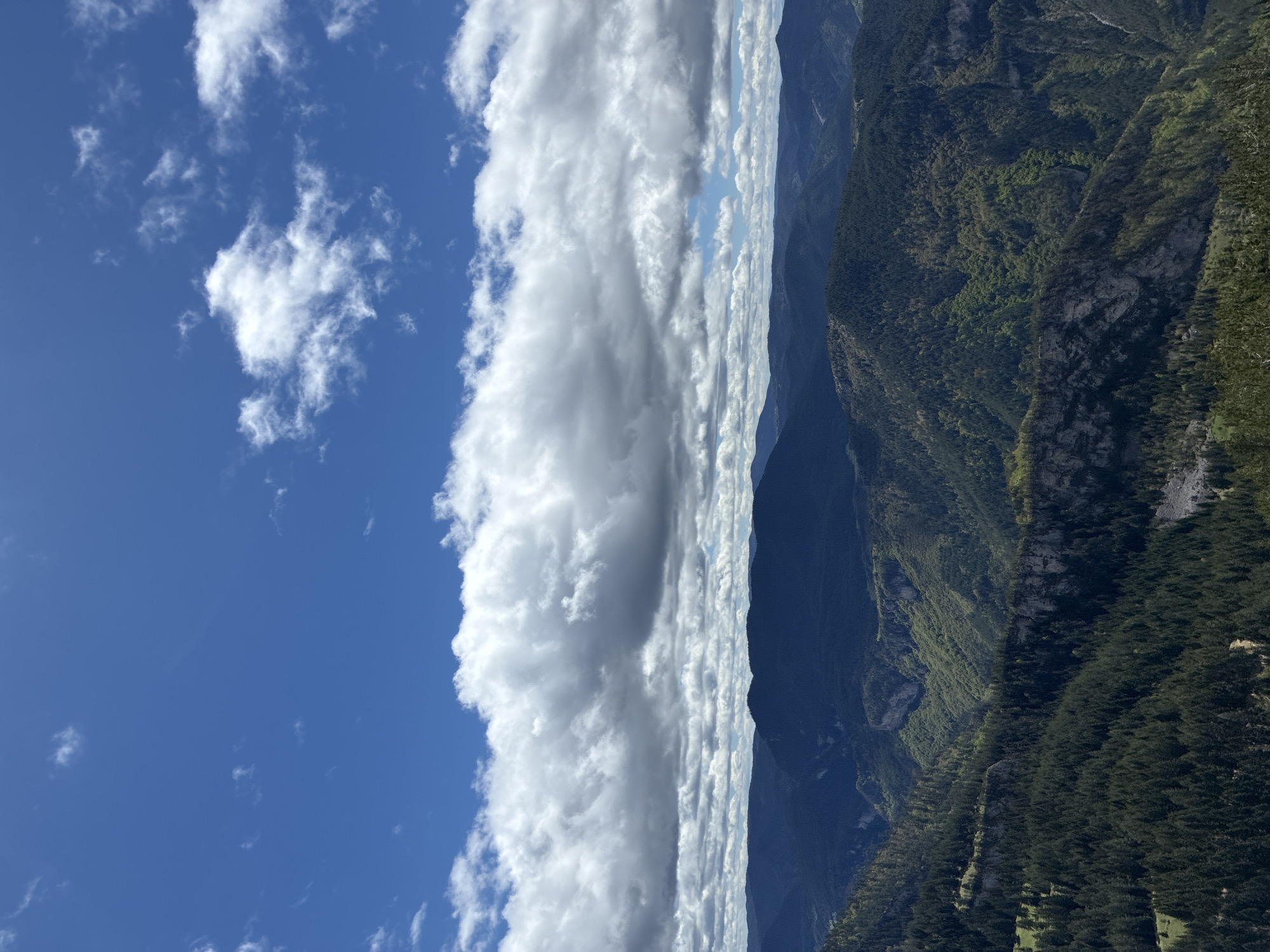 The image shows a landscape view with a blue sky, clouds, mountains, and forest. The sky occupies the left side of the image, featuring scattered white clouds. A thick band of white clouds stretches vertically down the center of the image. The mountains occupy the right side, with varying shades of green indicating forest cover. The location is likely Giovren, Bulgaria. There are no people visible in the image.