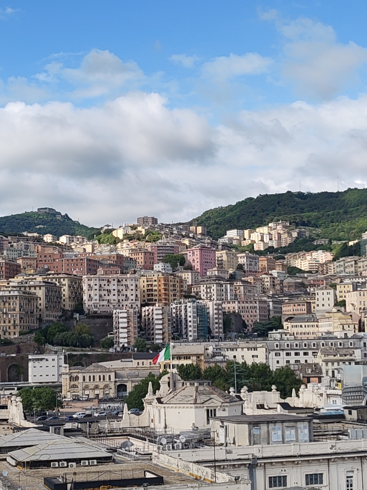 The image depicts a densely populated urban landscape in Genoa, Italy. In the foreground are rooftops and buildings with industrial or commercial characteristics. Moving toward the middle ground is a collection of buildings, diverse in architectural style and color, rising up a hillside. An Italian flag can be seen amongst these structures. In the background, a heavily forested mountain provides a natural backdrop. The sky is a mix of blue and puffy clouds, creating a bright, daytime setting.