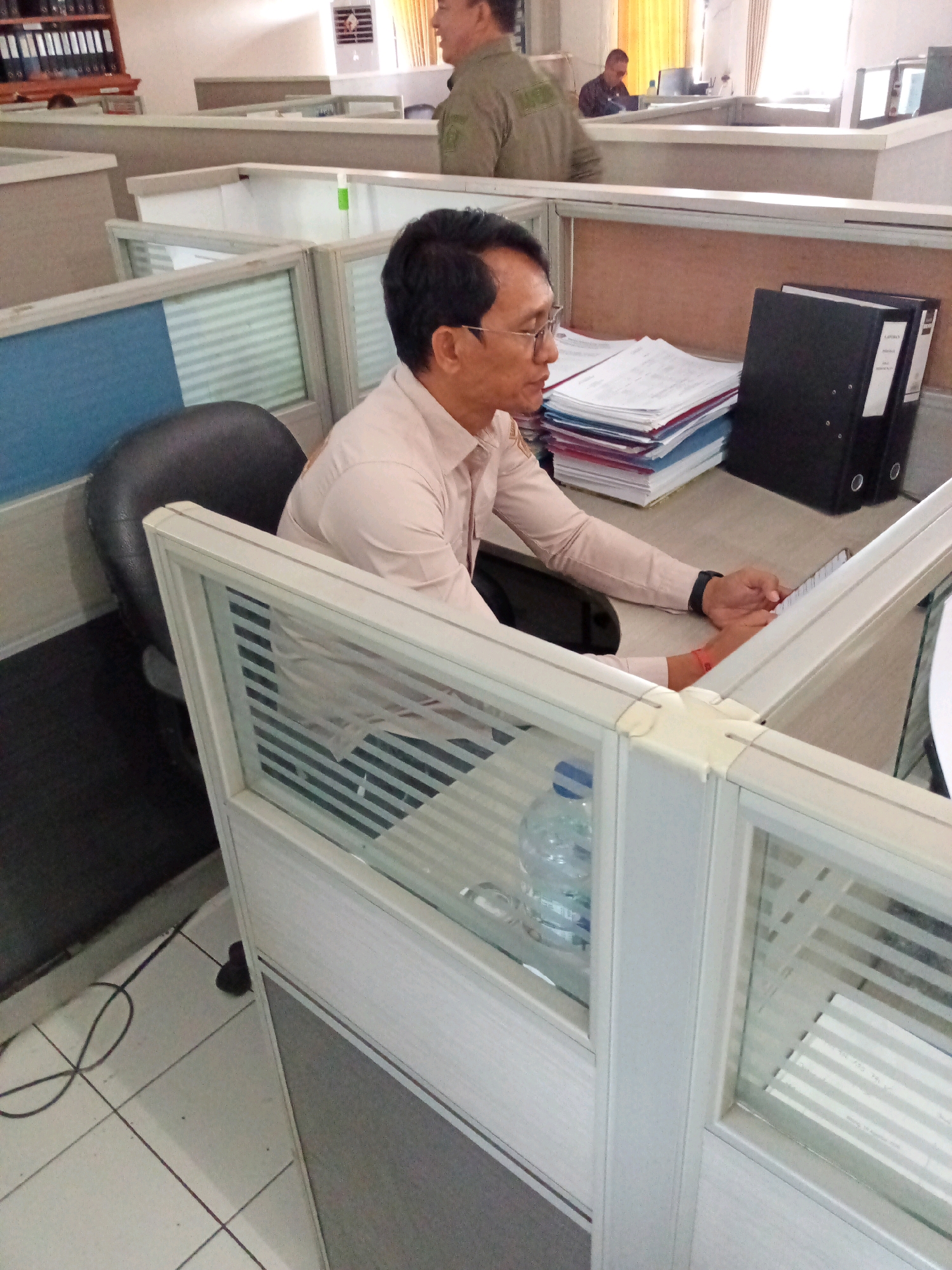 The image shows an indoor office environment with cubicles. In the foreground, a man wearing glasses and a light-colored collared shirt is seated at a desk, looking at a smartphone. His left hand is resting on the desk, and his right hand is holding the smartphone. To his left is a black office chair. On the desk, there are stacks of papers and two black binders. A partially full plastic water bottle is visible on a lower shelf. In the background, another person wearing a green uniform is standing. The office appears to be functional, with rows of cubicles, suggesting a typical workspace. The location in Bitung, Indonesia, is not visually identifiable from the image content.