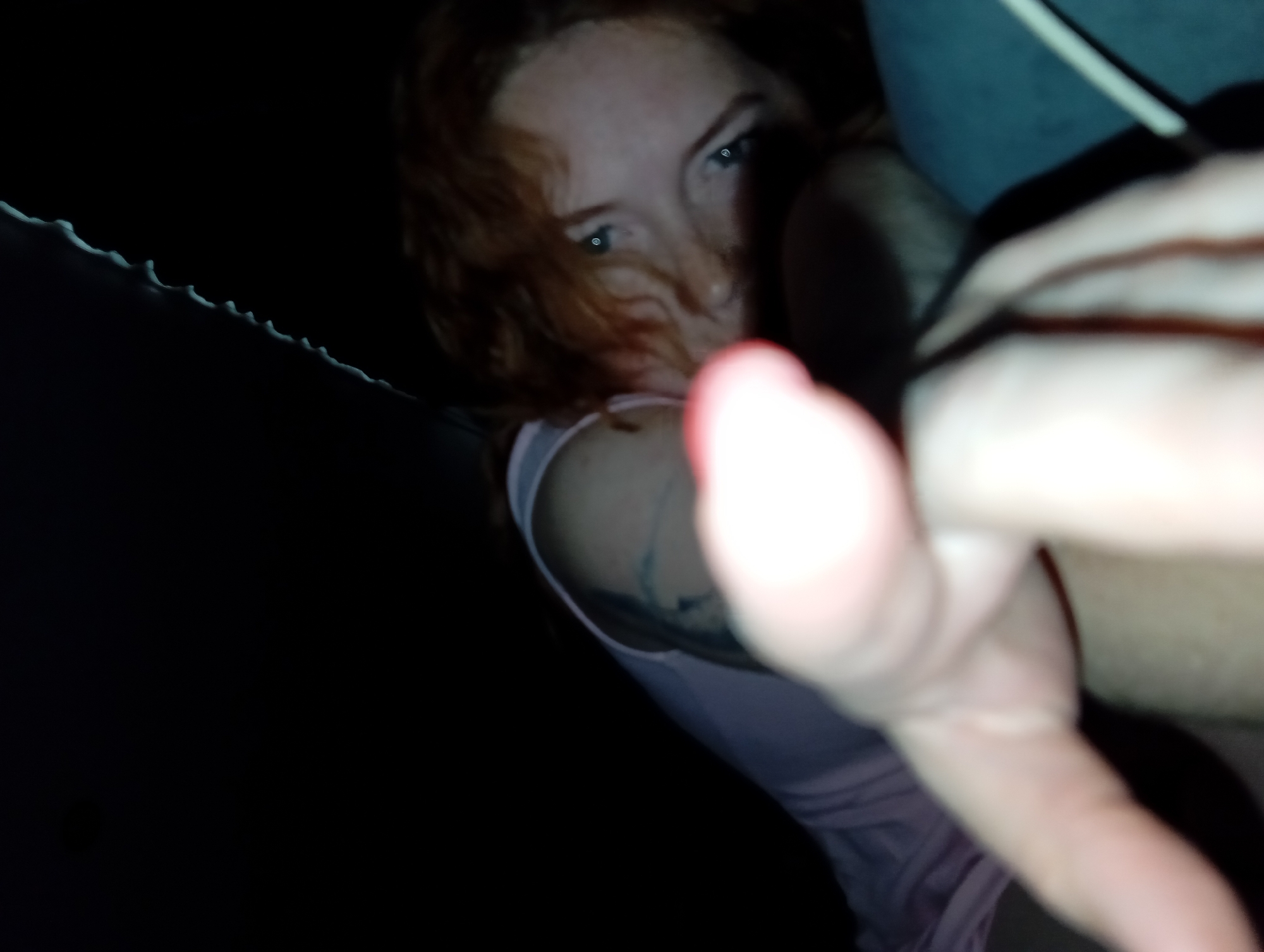 A low-angle photograph in a dark environment features a fair-skinned, red-haired individual with visible eyes gazing towards the camera. The person's right hand is extended towards the lens, obscuring the view. A tattoo is visible on their left shoulder. The individual is wearing a light pink top. A poorly lit background shows a possible ceiling. The location of the photograph is indeterminable, and there is no indication of being in Yurgamysh, Russia.
