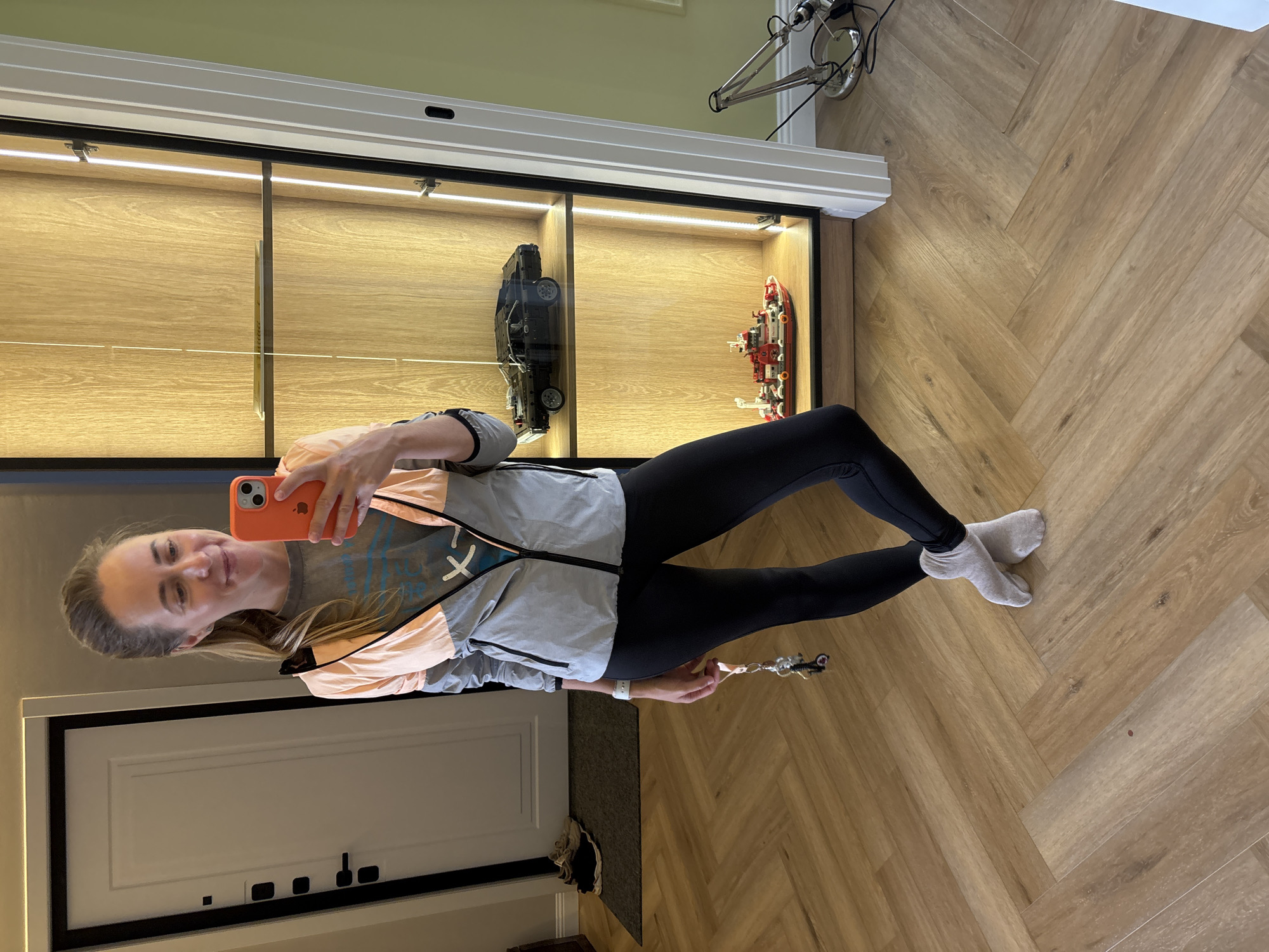 A woman stands in a hallway, taking a selfie in a mirror. She is wearing a grey and peach windbreaker jacket over a grey t-shirt, black leggings, and light grey socks. She holds an orange smartphone in her right hand, and a set of keys hangs from her left. A white smartwatch is visible on her left wrist. The hallway has light brown wood-plank flooring laid in a herringbone pattern. To the left of the woman is a white door with black trim and black door handles. A dark grey mat with two pairs of shoes is on the floor in front of the door. To the right of the woman is a tall, narrow display cabinet with glass doors, illuminated by warm LED lights. Inside the cabinet are two Lego models: a black car on the top shelf and a red and white boat on the bottom shelf. A silver desk lamp is positioned to the right of the display cabinet. The background wall is painted a light green. The image does not contain sufficient information to determine the location within the city of Chaikovsky, Russia.