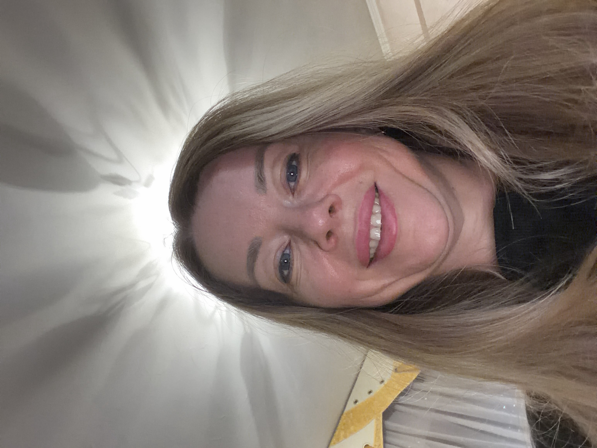 A selfie of a woman with blonde hair and blue eyes, smiling. She is wearing a black top. The background shows a ceiling with light and shadows, suggesting an indoor setting. There is a partially visible curtain and a decorative element on the left side of the image. The image does not provide any information about the location being Rome, Italy.