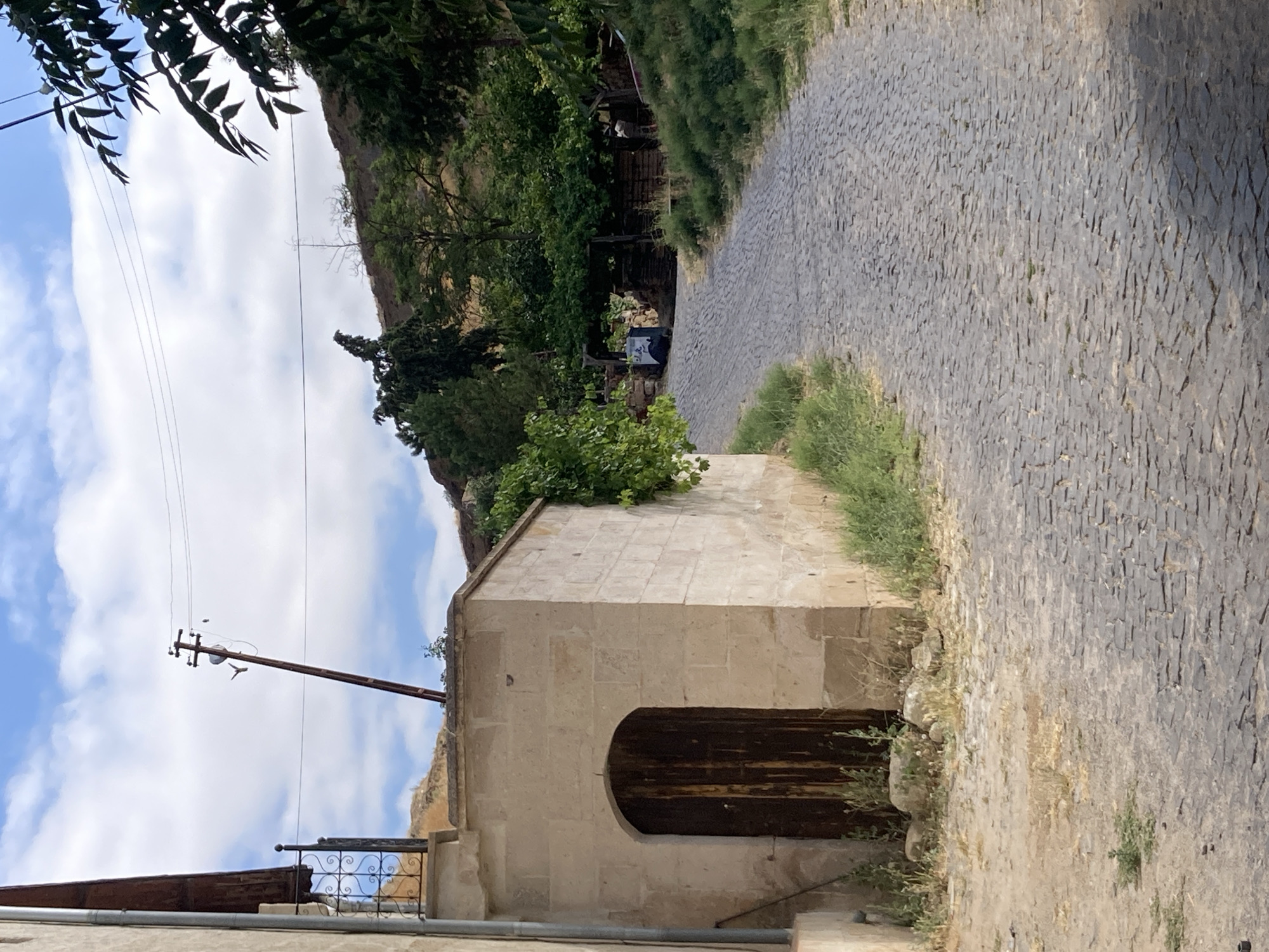 The image depicts a view of a narrow, winding cobblestone street in Avanos İlçe Merkezi, Turkey. The street is flanked by buildings constructed from light-colored stone. To the left, a building with a vertical downspout is partially visible, adjacent to a stone structure with a dark wooden arched doorway. This doorway is set into a light beige stone wall, with some patchy green vegetation growing around its base.
The cobblestone street itself is made up of irregular, gray stones, with some grass growing in the cracks. The street curves to the right and leads uphill. On the right side of the street, the terrain rises and is covered in dense green vegetation and trees.
In the background, a utility pole with wires is visible against a bright blue sky with scattered white clouds. The pole is leaning at an angle. Further up the hill, more trees and rocky terrain are present. A dark, possibly metal bin with some writing or symbols on it is situated near the base of the uphill vegetation. The overall scene suggests a historical or rural setting within the town.