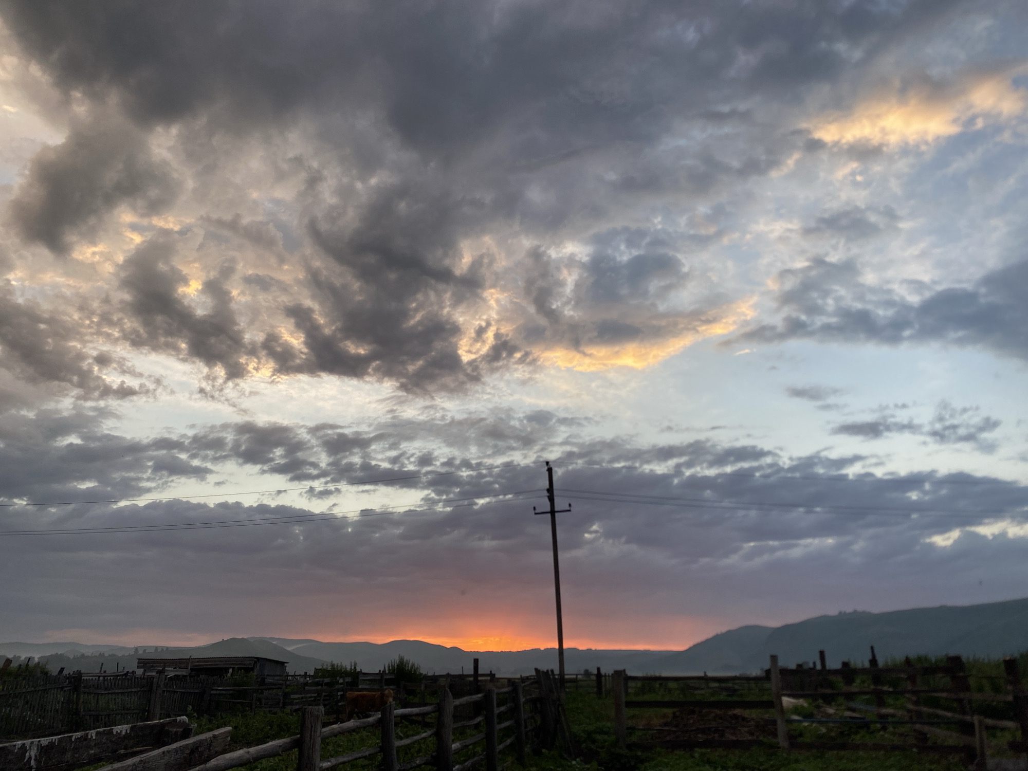 The image depicts a rural landscape at dusk, featuring a dramatic sky filled with clouds tinged with orange and yellow from the setting sun. In the foreground, a rustic wooden fence encloses a farm area, with some buildings and a solitary cow visible within the enclosure. A single utility pole stands prominently in the middle ground. The background shows a silhouette of rolling hills, suggesting a mountainous or hilly terrain. The scene is set in the village of Maloarslangulovo, Russia.