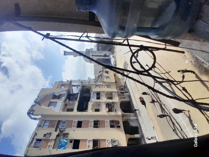 The image depicts a damaged urban street scene in Khan Yunis, Palestinian Territory, viewed from an elevated perspective, likely a window or balcony. In the foreground, a large clear plastic water bottle on the right and a network of dark wires, rods, and a circular metal object partially obstruct the view.
The central feature is a multi-story building on the left with significant structural damage, including partially collapsed upper floors, exposed interiors, and missing sections of walls. A blue tarp is visible in one window opening on the lower-left side of this building. Across the street, another building with multiple balconies also shows signs of damage.
In the mid-ground, a street runs horizontally across the frame. Approximately 10-15 individuals, dressed in civilian clothing, are dispersed along the street. Some are walking individually, while a smaller group of 3-5 people appears to be sitting or gathered on the left side of the street. Shadows from the people and foreground obstructions are cast on the road.
In the background, a large mosque or religious structure with two prominent minarets and a central dome is visible. This structure also appears to have sustained damage, with rubble visible at its base. The sky above is partly cloudy with patches of blue. The overall environment suggests a heavily damaged urban area. A "Galaxy A14" watermark is present in the bottom left corner.