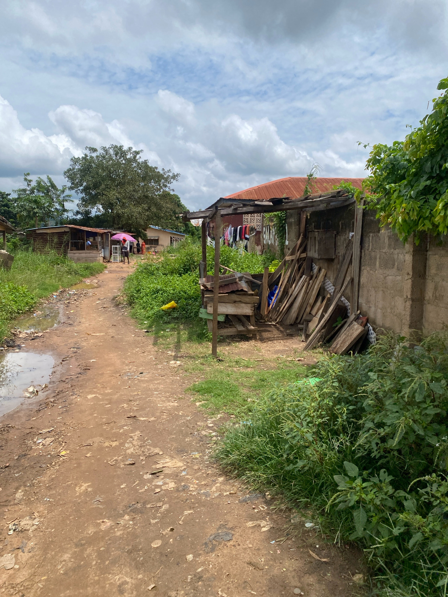 The image depicts a dirt road in a neighborhood in Iworoko, Nigeria. The road is uneven and appears to be wet in places, with puddles of water and scattered debris. On the right side of the road, there is a concrete wall and a makeshift shelter constructed from wood and corrugated metal. This shelter is filled with stacks of lumber. Clothing hangs on a clothesline near another structure with a rusted metal roof in the background. To the left of the road, there are more small structures, one with a pink umbrella over a person standing outside. Dense green vegetation grows along the sides of the road and over some of the structures. The sky is partly cloudy with a mix of white clouds and blue sky.