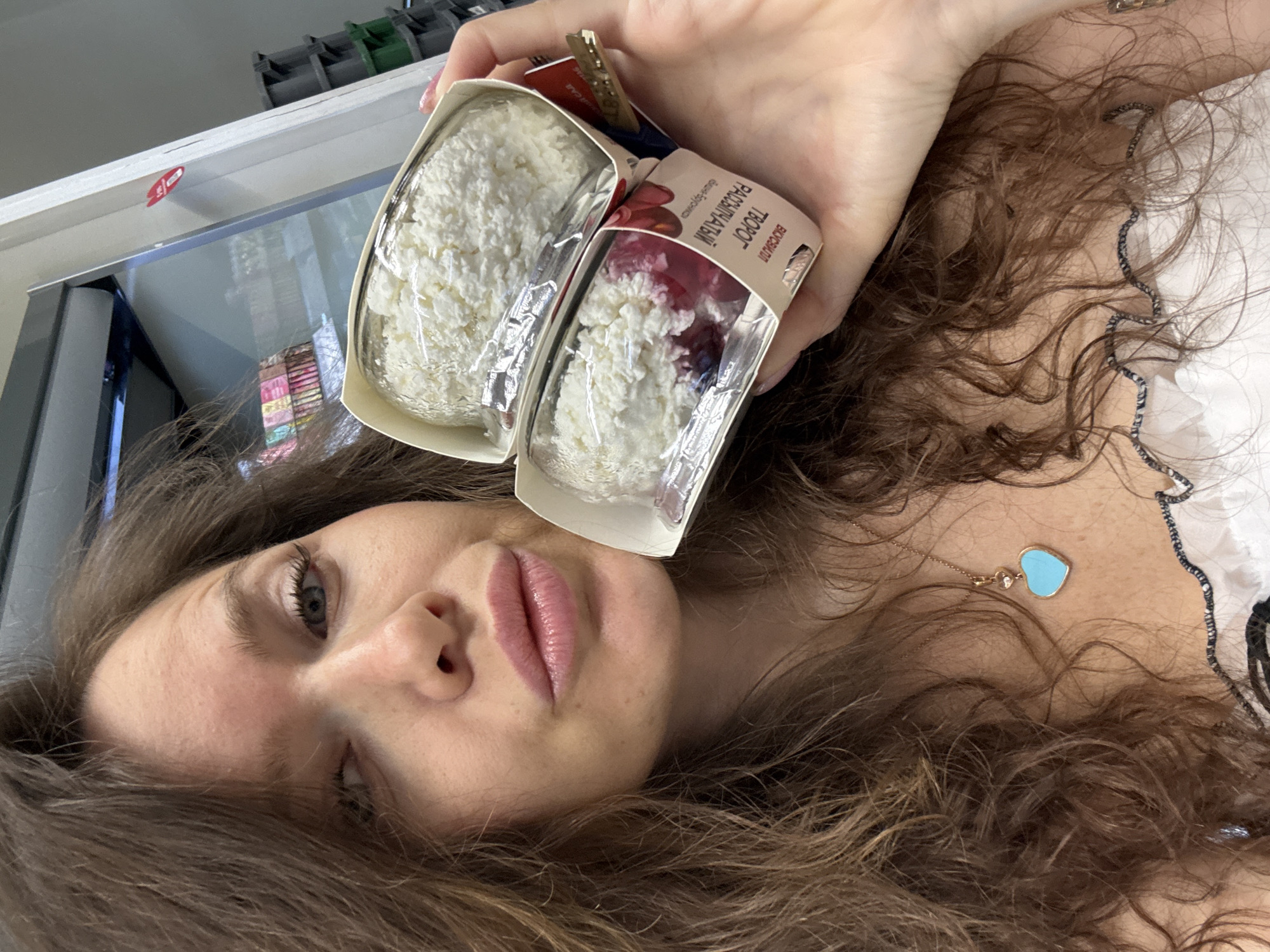 A close-up, slightly angled selfie captures a woman holding two containers of cottage cheese. The woman, with long, wavy brown hair, looks towards the viewer. Her left eye is visible, and her lips are slightly parted, adorned with light pink lip color. She wears a light-colored top with some dark trim and a delicate gold-toned necklace featuring a small turquoise heart-shaped pendant and a clear gem.
She holds two clear plastic containers of cottage cheese, presented in a cardboard sleeve. The left container holds plain, crumbly white cottage cheese. The right container features similar cottage cheese topped with a layer of red fruit or berry jam. Visible text on the packaging is in Cyrillic, identifying the product as "ТВОРОГ РАССЫПЧАТЫЙ" (Crumblу Cottage Cheese) and displaying the brand name "ВКУСНОТЕЕВО" (Vkusnoteevo).
The background, partially out of focus, suggests an indoor setting. Reflections of shelves or other items are visible in what appears to be a glass surface or display case, possibly in a store. The lighting indicates it is daytime. The woman's expression is neutral. The Russian text on the product packaging aligns with the context of Khimki, Russia.
