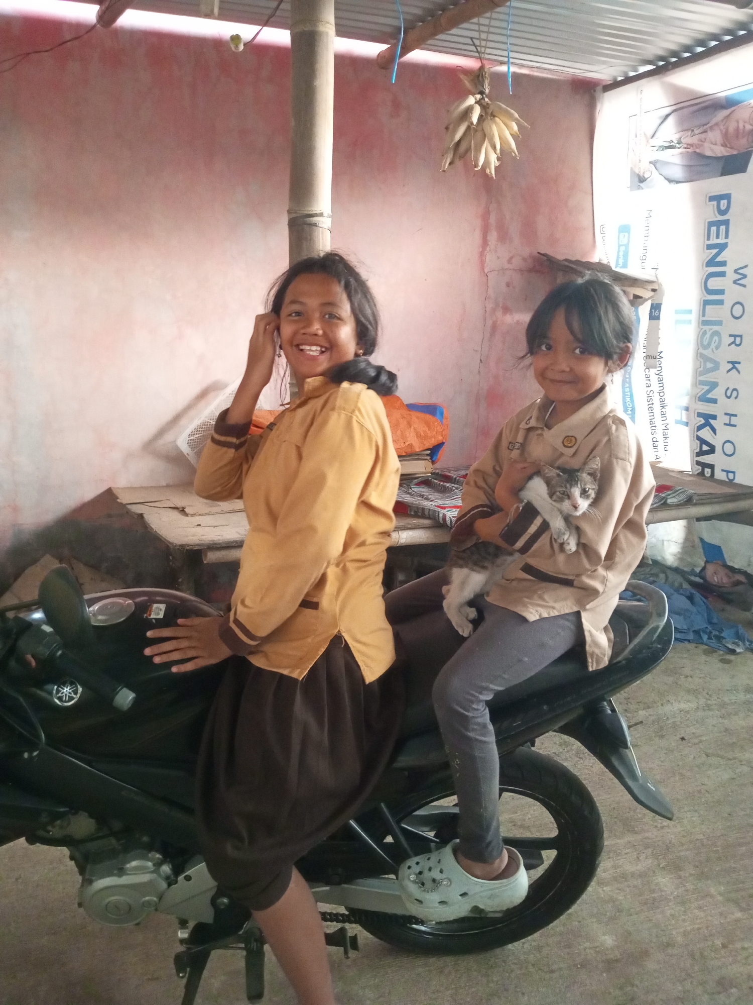 In Andongsili, Indonesia, two young girls, likely students based on their school uniforms, are photographed sitting on a black motorcycle. The girl on the left, wearing a tan uniform with brown trim, is smiling and touching her ear. The girl on the right, also in a tan uniform with brown trim, is holding a small tabby cat. Both girls are wearing dark pants and casual shoes. The motorcycle is parked on a concrete floor. The background consists of a pinkish-red wall, a wooden table, and a corrugated metal roof. A bunch of dried corn hangs from the ceiling, and a poster is visible on the right wall. The environment appears to be a modest, possibly rural, dwelling or community space.