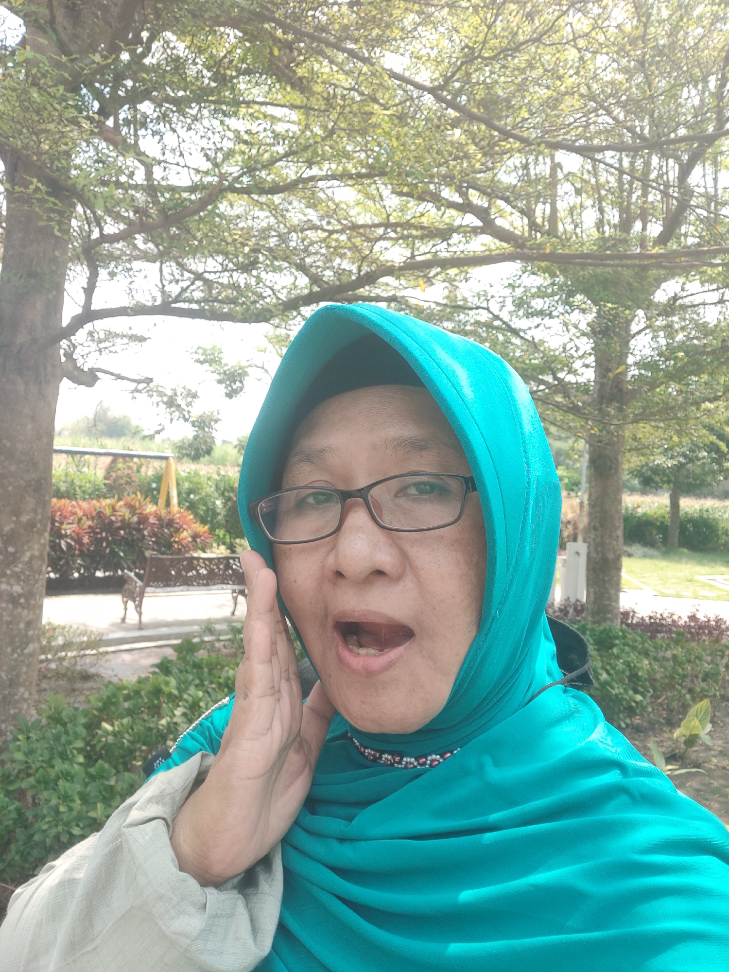 A middle-aged woman wearing a teal hijab and glasses is posing for a selfie in an outdoor park in Pagu, Indonesia. She is holding her right hand to her mouth and her mouth is open, as if speaking or shouting. The background shows trees, greenery, and a bench.