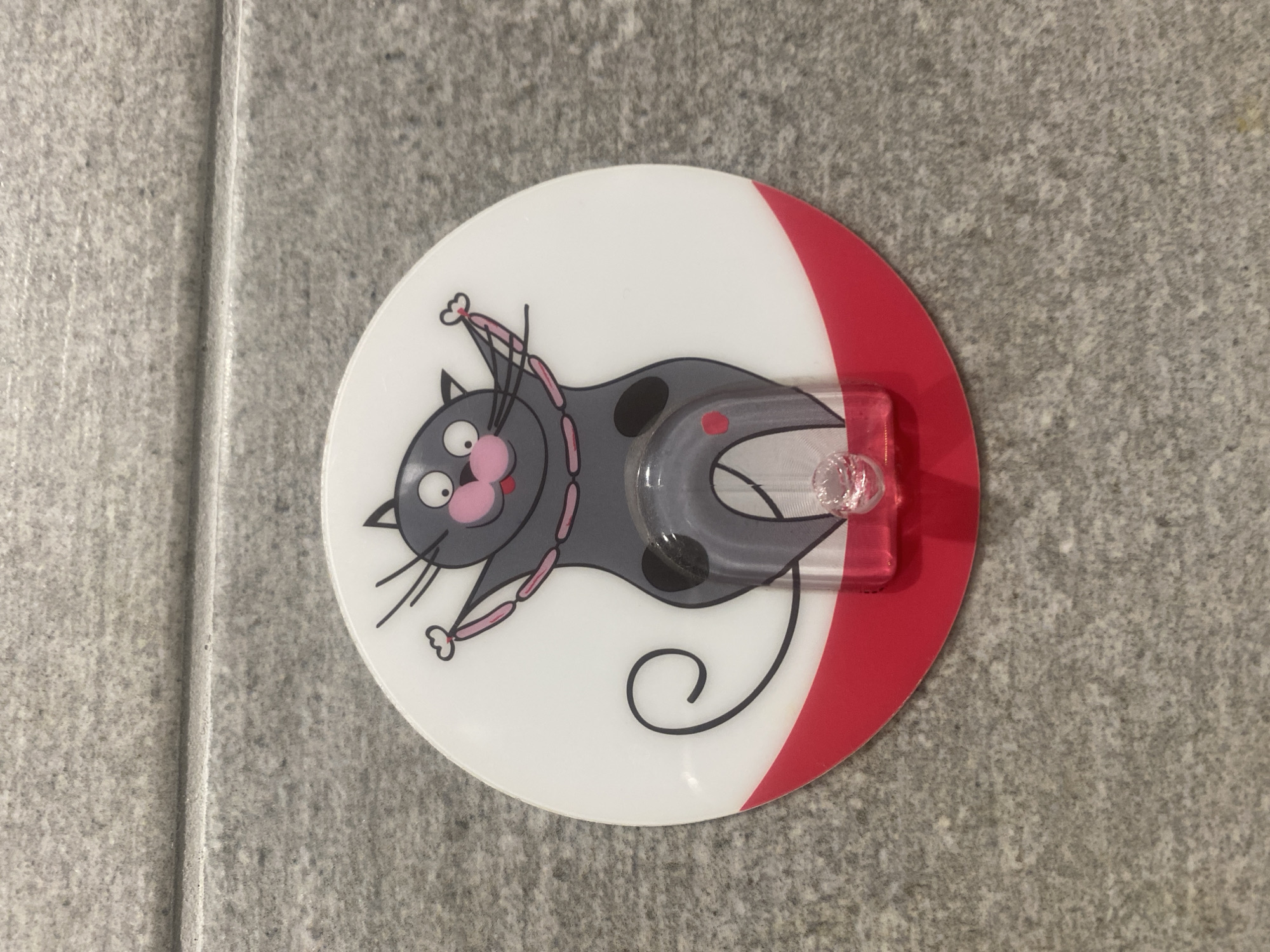 A round, white wall hook with a cartoon image of a gray cat is affixed to a gray tiled wall. The cat has pink cheeks, a pink nose, large eyes, and is wearing a pink beaded necklace. Its arms are raised in a welcoming gesture, and its tail curves around its body. A clear plastic hook extends from the cat's midsection. The bottom of the hook is decorated with a red semi-circle. The wall appears to be made of speckled, light gray tiles with visible grout lines. The image does not provide any information about its location in Chișinău, Moldova.