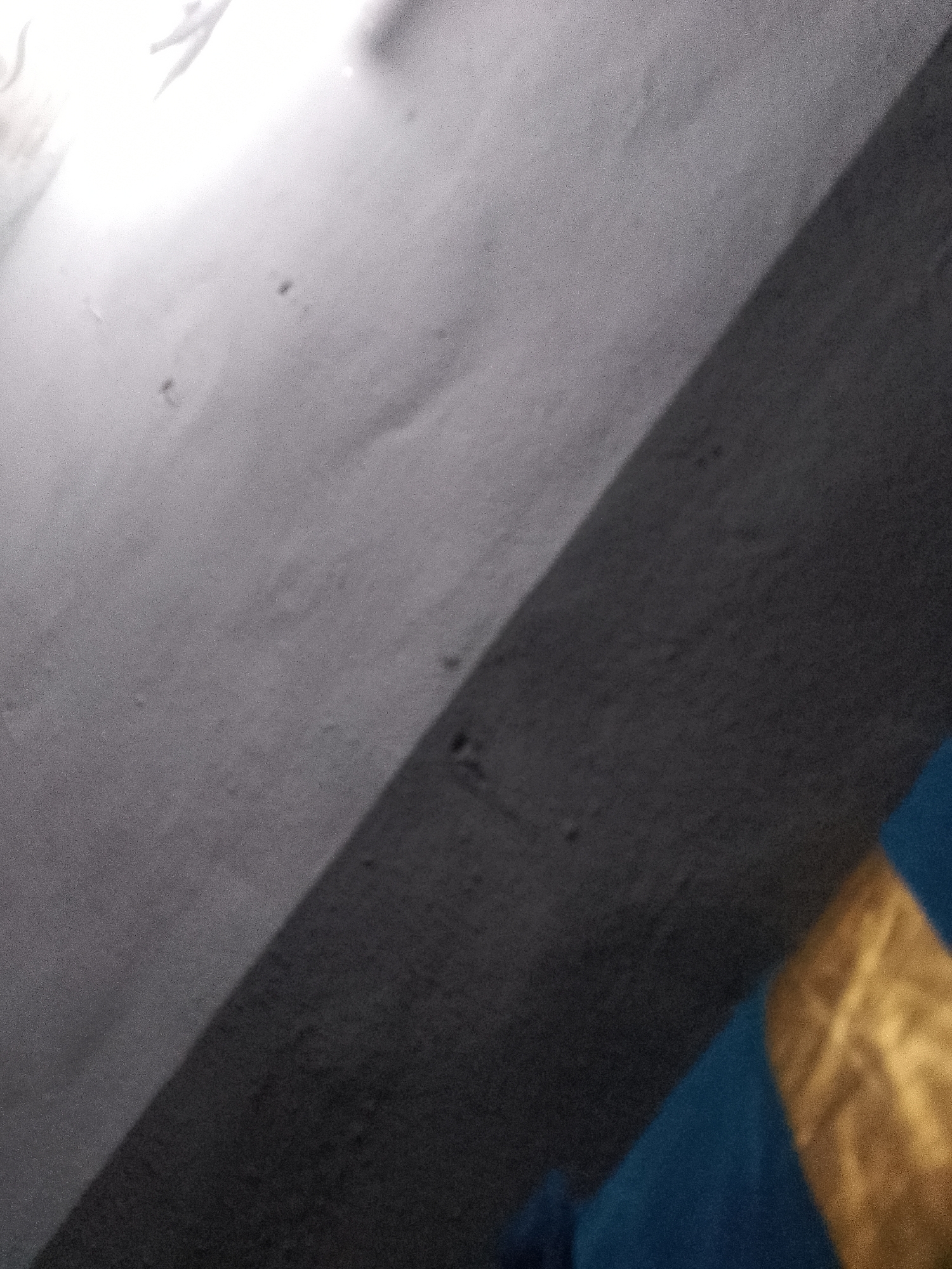 The image depicts a textured surface, likely a wall, with a distinct diagonal shadow line dividing it into a lighter upper portion and a darker lower portion. The lighter section appears to have some subtle imperfections or markings. In the bottom right of the frame, a portion of what appears to be fabric or clothing is visible, predominantly blue with a golden, patterned band. The overall lighting suggests an indoor or shaded outdoor environment. The image lacks sufficient context to determine its specific location within Jauro Yinu, Nigeria, or to identify any people or specific actions.