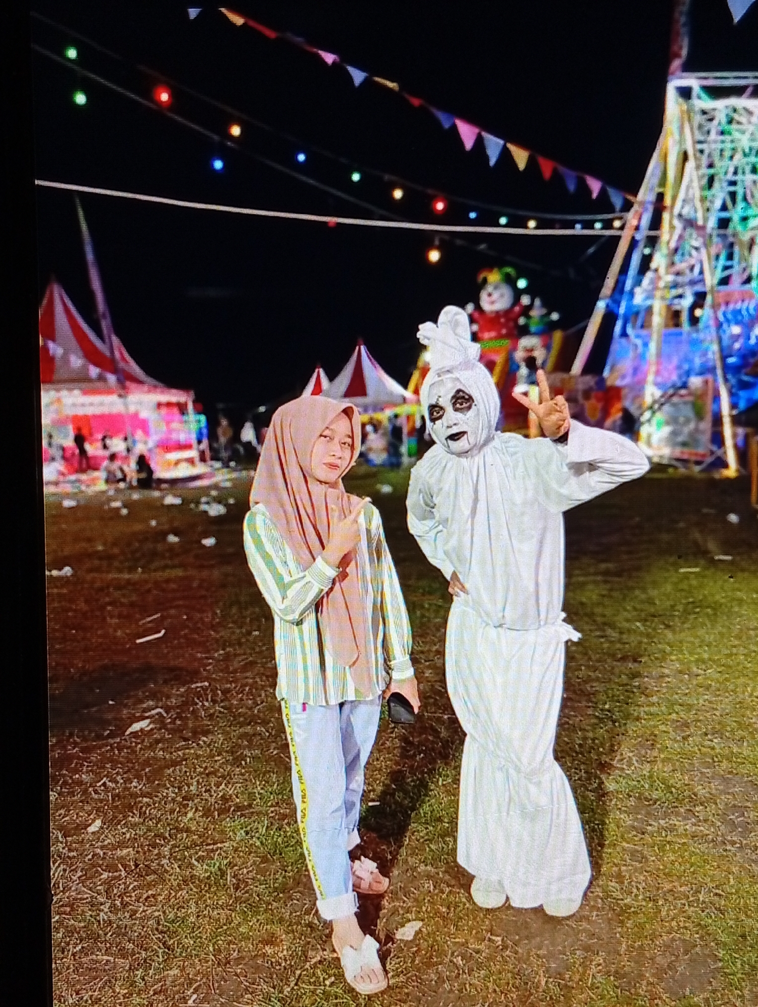 This image depicts two individuals standing outdoors at night in an event space located in Lhokseumawe, Indonesia. The ground is a mixture of sparse grass and dirt.
On the left, a female individual with light skin wears a light brown hijab, a vertically striped long-sleeved shirt (featuring green, white, and brown stripes), light blue pants with a yellow 'FILA' brand stripe on the outer seam, and light-colored sandals. She holds a dark-colored smartphone in her left hand and extends her right arm with the index finger pointing forward. She is looking directly at the camera.
On the right, an individual is dressed in a full white costume resembling a ghost. The head is wrapped in white fabric, secured at the top, and the face is painted with black around the eyes and a black mouth. The costume consists of a white long-sleeved top and loose white bottoms. This individual wears light-colored footwear, places the left hand on the hip, and raises the right hand in a 'V' gesture. This individual is also looking directly at the camera.
In the background on the left, multiple tents with red and white striped roofs are visible, illuminated by various lights, including pink and purple hues. Several people are discernible around and within these tents. Towards the center-right background, a tall, brightly lit structure, possibly an amusement ride like a Ferris wheel, displays multi-colored lights (blue, green, pink). Behind the costumed individual, a large inflatable figure, appearing to be a clown or a similar character with a red body and colorful head, stands as part of another attraction. Spanning the upper portion of the background are string lights with multi-colored bulbs (green, red, orange, blue) and decorative triangular bunting flags in various colors. The overall scene is indicative of a night fair or carnival environment.