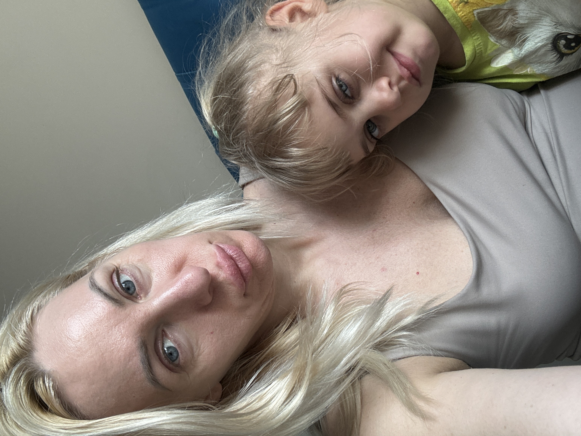A close-up shot depicts a woman and a young child lying down. The woman has blonde hair and blue eyes and is wearing a beige top. A young girl with blonde hair is next to the woman. She is smiling at the camera. She is wearing a shirt with an animal on it. The environment appears to be indoors, with a blue headboard visible in the background. The location is Unknown, Unknown.