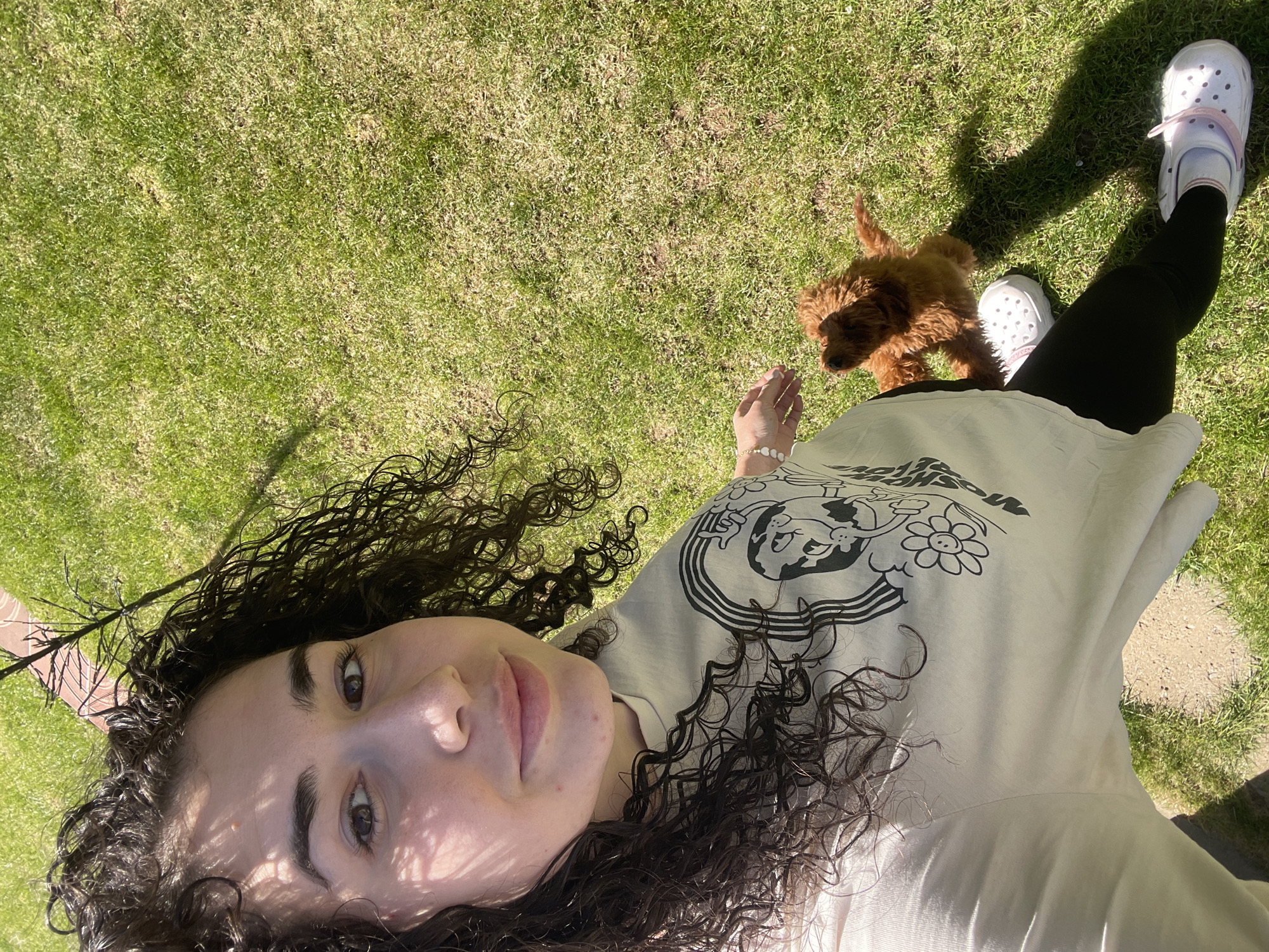 The image depicts a serene outdoor scene in Sofia, Bulgaria, featuring a young person and a small puppy on a sunny day. The camera is oriented as if looking down from above, with the subjects lying on a green lawn.
The main subject is a person with dark, curly hair, lying on their side with their head turned towards the camera. Their face is partially shaded by dappled light, revealing dark eyes and a neutral expression. They are wearing a light-colored, possibly cream or beige, T-shirt with a graphic design featuring cartoon-like figures, including a smiling flower, and some indiscernible text. Black leggings cover their legs, and white Crocs-style shoes with light pink straps are visible on their feet. A white beaded bracelet adorns their right wrist.
Adjacent to the person's leg stands a small, light brown, fluffy puppy, with curly fur, looking towards the person's hand. The puppy appears to be a Poodle mix.
The setting is a vibrant green lawn, bathed in bright sunlight, which casts strong, clear shadows from both the person and other unseen objects, suggesting it is midday or early afternoon. The grass appears well-maintained. A faint outline of a paved path or brick border is visible in the background. The overall atmosphere is relaxed and peaceful, capturing a tender moment between the person and their pet.