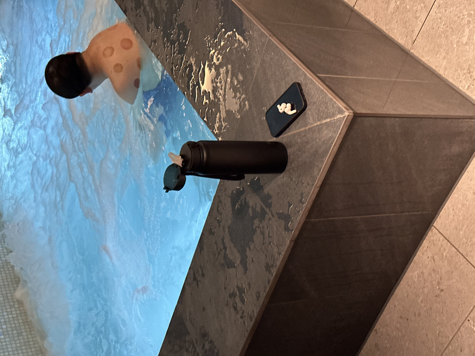 This image depicts an indoor scene at what appears to be a spa, gym, or wellness center in London, United Kingdom. A person with dark hair is partially submerged in a light blue body of water, likely a hot tub or small pool, facing away from the camera. Numerous distinct, circular red marks, indicative of cupping therapy, are visible across their upper back. The water around them shows signs of agitation, consistent with a jacuzzi or similar feature.
On the dark grey tiled edge beside the water, a black water bottle is placed horizontally with its lid open, revealing a spout. Next to the bottle, a black smartphone with white wireless earpods resting on its screen is visible. The surrounding tiled surfaces are wet in places, suggesting proximity to the water. The overall lighting is artificial, characteristic of an indoor facility, making the specific time of day indeterminate. No discernible text is visible in the image. The scene suggests a moment of relaxation or recovery after a therapeutic treatment.