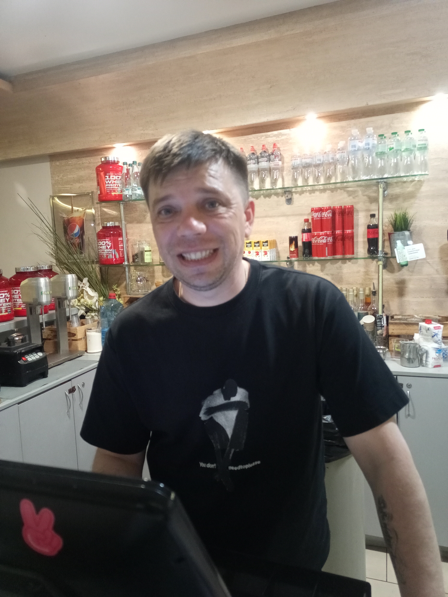 This media file is a medium shot photo of a man standing behind a counter. The man, who appears to be in his late 30s or early 40s, has short brown hair and is smiling directly at the camera. He is wearing a black t-shirt with a white graphic and some text that is partially visible.
The background of the image suggests a retail environment, possibly a health supplement store or a cafe. Shelves behind the man are stocked with various items. On the left, there are red tubs labeled "100% WHEY PROTEIN," and a Pepsi display. Further back, there are rows of bottled drinks, including Coca-Cola cans and bottles of water. There are also some kitchen appliances, such as blenders and milkshake machines, on the counter.
The lighting is bright, indicating it might be daytime. The overall mood of the photo is casual and friendly, conveyed by the man's smile. The text on the man's shirt reads "You don't needtopludse," and the Coca-Cola cans are clearly visible in the background. The presence of fitness supplements and beverages suggests a business focused on health and refreshment.