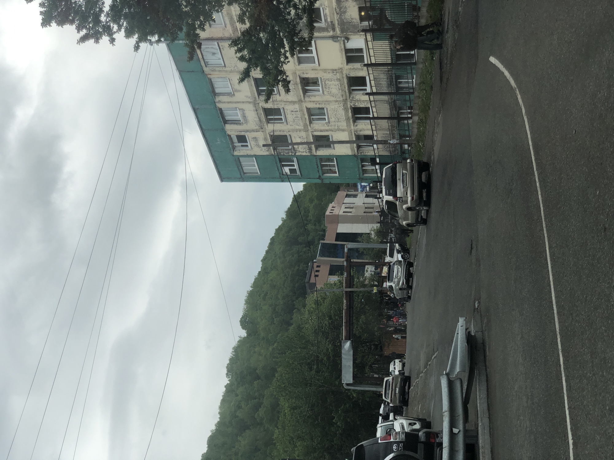The image depicts a street scene in Petropavlovsk-Kamchatsky, Russia, under an overcast sky. In the foreground, a grey asphalt road curves to the right with a white line marking its edge. A bent metal guardrail is visible on the left. Several vehicles are parked or driving on the road, including a dark SUV on the far left, a silver SUV, a white car, and another silver SUV further down the road.
To the right of the road is a multi-story building with a faded yellow facade and a teal-colored upper section. The building has numerous windows, many of which appear to be covered with curtains or blinds. A black metal fence runs along the front of the building. Two individuals, dressed in dark clothing, are standing near the fence.
On the left side of the image, a steep, tree-covered hillside dominates the background. In the middle ground, between the road and the hillside, there are other buildings, including one with a modern facade featuring large dark windows. A metal structure, possibly a gantry or support for overhead wires, is visible in front of these buildings. A group of people are gathered on the sidewalk or near the road in front of these buildings. Power lines are strung across the upper portion of the image.