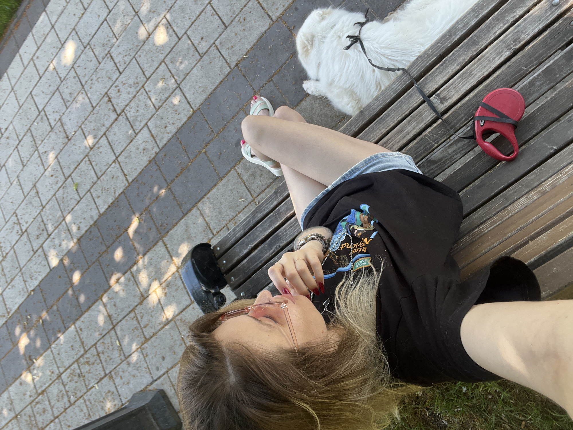 A young woman with blonde hair, wearing pink sunglasses, a black t-shirt with a colorful graphic, and denim shorts, sits on a wooden park bench with her legs crossed. A white fluffy dog lies on the bench next to her, attached to a red retractable leash. The bench is located on a paved pathway with square and rectangular grey and brown tiles, with dappled sunlight shining through. The scene appears to be outdoors, possibly in a park or public walkway.