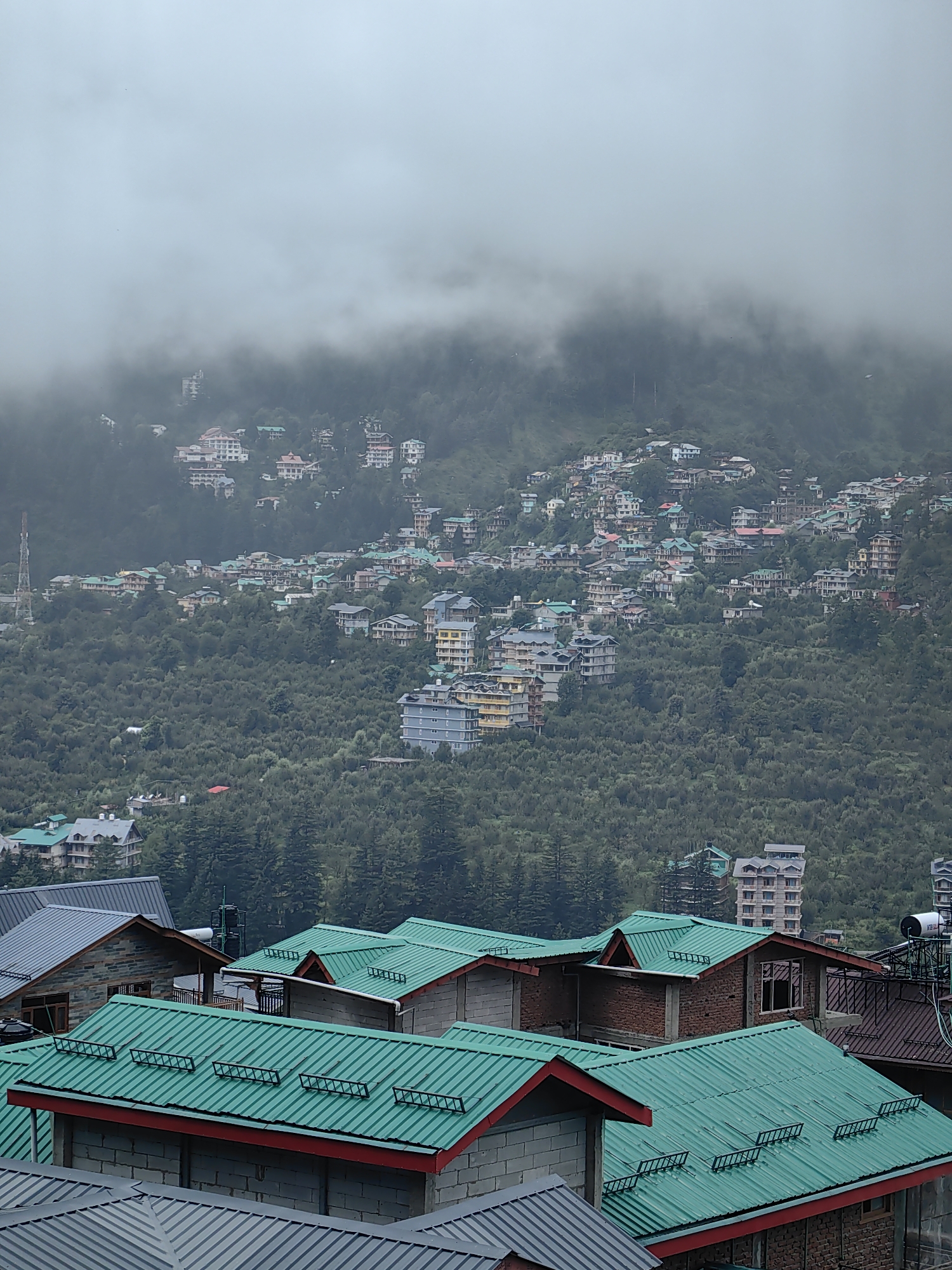 This is a vertical, eye-level, full shot of a hillside town shrouded in thick clouds. The image is dominated by the dense green foliage of a mountainside, upon which a multitude of buildings are nestled. The architecture varies, with many structures featuring brightly colored roofs, predominantly green and red. Some buildings are multi-story with balconies, while others are smaller, more traditional homes. The scene is characterized by a misty, atmospheric quality due to the heavy cloud cover that obscures the upper reaches of the mountain and the sky. The foreground shows several buildings with distinct green metal roofs, some with red trim, and brick or concrete walls. In the midground and background, the town extends up the hillside, with buildings becoming smaller and less distinct as they recede into the mist. A cell tower is visible to the left in the midground. The overall impression is one of a densely populated hillside settlement in a mountainous region, possibly Vashisht, India, given its known characteristic as a popular tourist destination with similar architecture and topography.