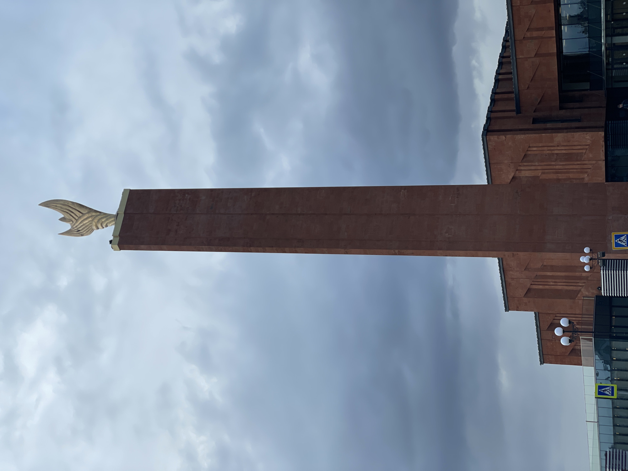 The image displays a tall, narrow, brown tower or obelisk topped with a golden, stylized, wing-like or fin-like sculpture, set against a cloudy, overcast sky. Adjacent to the left side of the tower stands a multi-story building constructed with brown or terracotta-colored materials, featuring a modern architectural design. This building includes large sections of dark, reflective glass windows that mirror the sky, and its facade has layered or stepped elements.
Several details are visible on the building: decorative lampposts with multiple spherical white lights are mounted on its exterior. Two blue square signs, each displaying a white triangle with a black pedestrian silhouette (pedestrian crossing signs), are affixed to the building's facade, one clearly visible in the lower right corner and another higher up.
The scene suggests an urban or monumental outdoor setting under daytime conditions with a heavily clouded sky. No human activity or specific events are depicted, drawing attention to the architectural structures themselves. The perspective is angled upwards, emphasizing the height and scale of the tower and building.