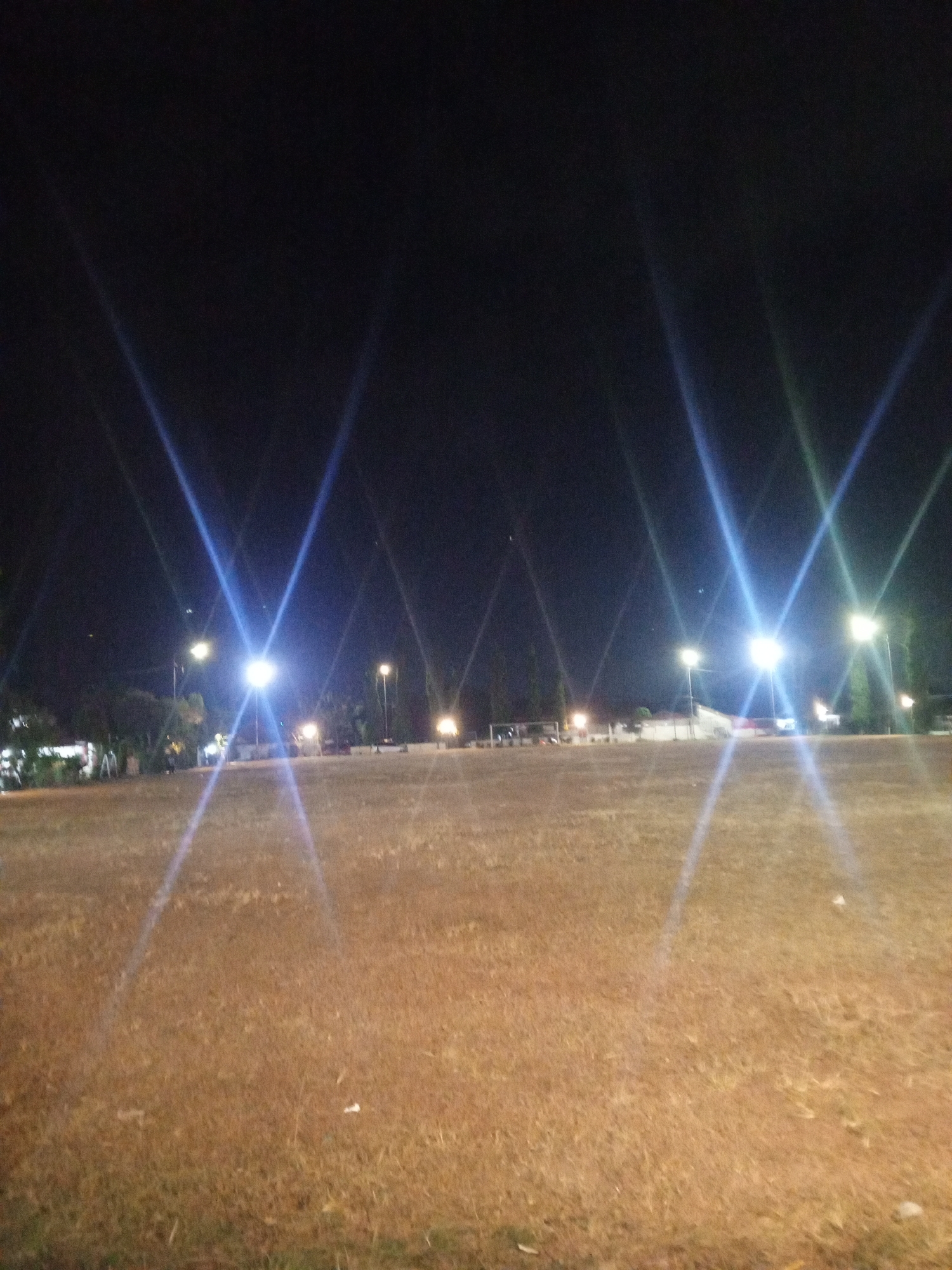 The image depicts an outdoor scene at night in the city of Kloncing, Indonesia. The foreground is dominated by a large, dry, grassy field, appearing somewhat barren. In the background, several tall light poles illuminate the area with bright, lens-flared lights, creating strong beams of light that extend across the frame. Scattered trees and some structures, possibly buildings or fencing, are visible behind the field. The overall impression is of an open space, possibly a park or a sports field, at night, with artificial lighting creating a dramatic effect. There are no identifiable people or specific objects of interest within the frame, apart from the lighting fixtures and the natural elements.