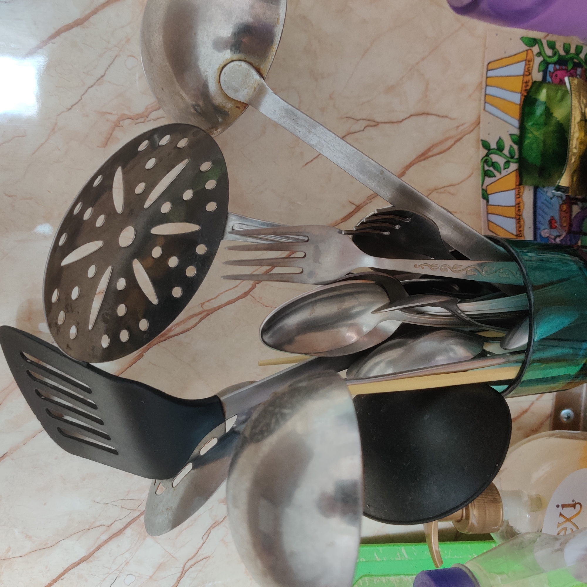 The image shows a close-up of a collection of kitchen utensils, including spatulas, ladles, spoons, and a fork, arranged in a green glass. The utensils are made of metal and black plastic. Behind the utensils, a wall with a marble-like pattern is visible. To the left, a bottle of liquid soap with a pump dispenser is partially visible, along with a green plastic item. To the right, a decorative tile with images of lamps and plants is present, and a portion of a purple object is seen at the edge of the frame. The scene appears to be a kitchen counter. There is no information in the image that would indicate the location in the city of Yekaterinburg, Russia.