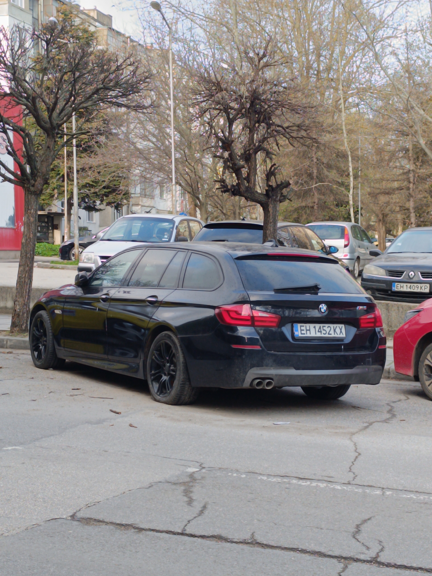 The image captures a black BMW station wagon parked on a cracked asphalt surface. The car, bearing a Bulgarian license plate "EH 1452 KX," is flanked by other vehicles, including a red car to the right and several cars in the background. The setting appears to be a city street, with bare trees lining the road and buildings visible in the background. The time of day seems to be daytime, under overcast lighting conditions. The city is Unknown, Unknown.