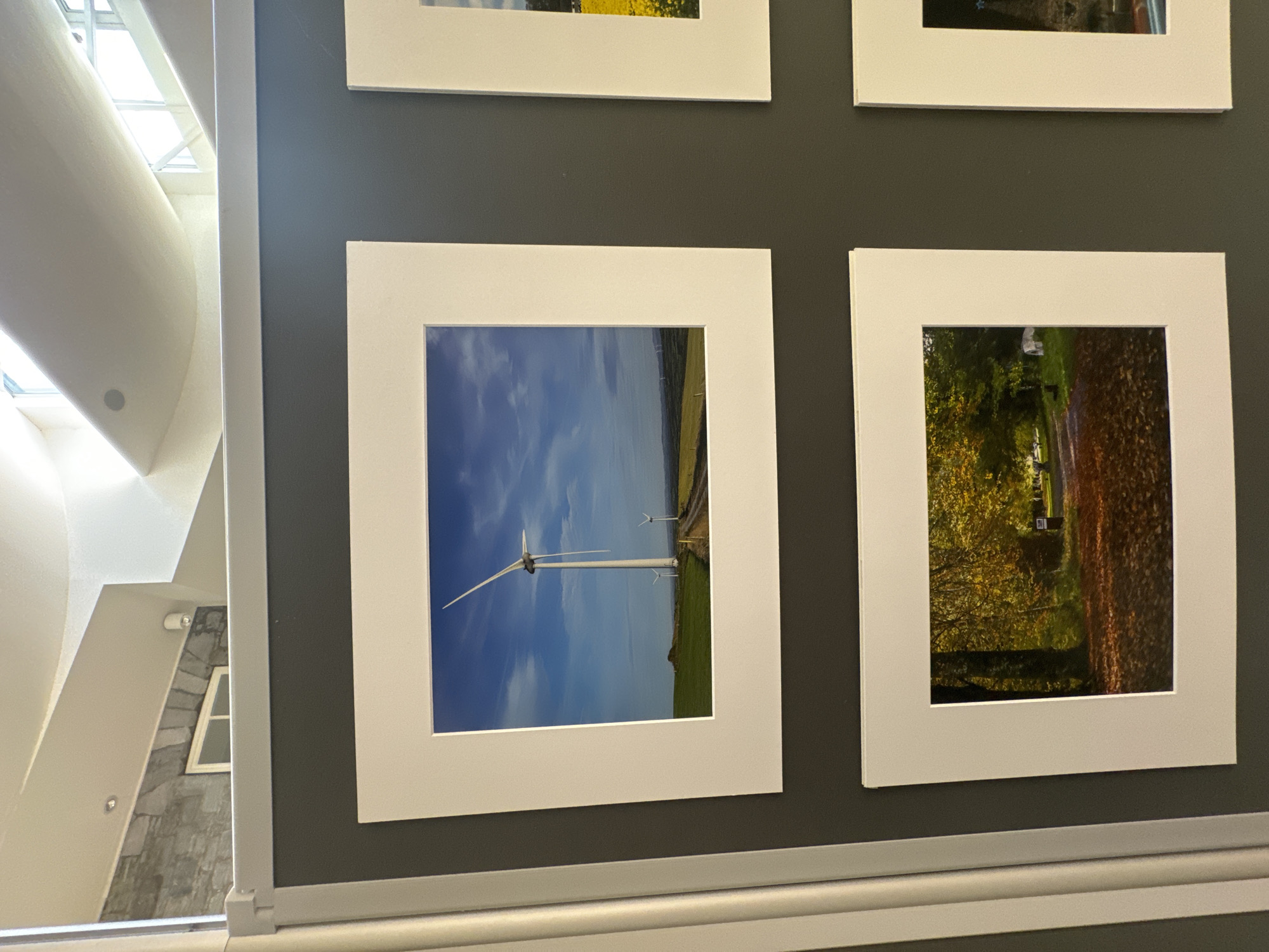 The image displays a grid of framed photographs mounted on a dark grey wall, likely in a gallery or exhibition space in Kilkenny, Ireland. The top row features two landscape photographs. The left photograph shows a wind turbine in a green field under a blue sky with scattered clouds. In the background, two more wind turbines are visible, along with a dirt road. The right photograph, partially visible, appears to be a field of yellow flowers. The bottom row also contains two photographs. The left photograph captures an autumn scene with a path covered in fallen leaves, bordered by trees with yellow and orange foliage. The right photograph is partially visible and seems to show a Christmas tree. The overall setting suggests an indoor exhibition.