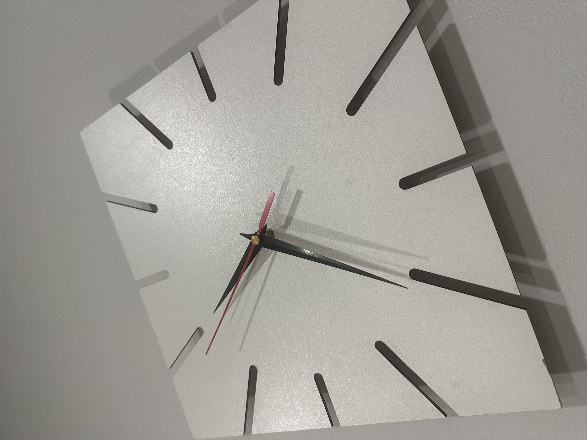 A square white wall clock hangs at an angle on a light gray wall. The clock face has dark gray markings for the hours, instead of numerals. The hour hand is red and points between the 7 and 8. The minute hand is black and points at the 1. The second hand is a thin pink needle. The time appears to be around 7:06 AM/PM. The wall is featureless, and there is nothing to suggest the location is Sochi, Russia.