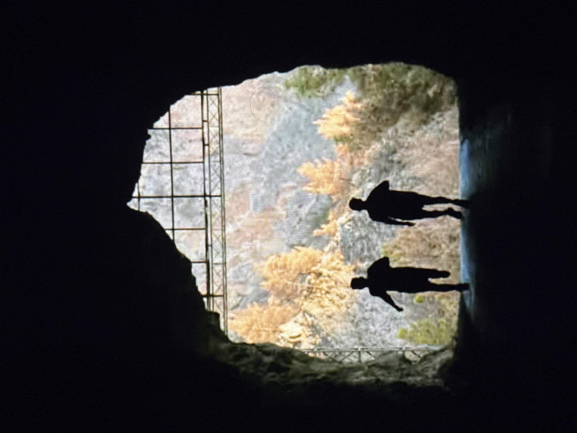 The image shows a dark, possibly cave-like structure framing a view of an outdoor area. Through the opening, a landscape is visible, featuring what appears to be a hillside with trees, some of which exhibit autumn colors. Two dark silhouettes resembling human figures are positioned in the opening, appearing to be in a horizontal orientation, possibly floating or jumping. To the left of the opening, a grid-like structure resembling scaffolding is partially visible. The image appears to be taken from a darkened interior, with the focus on the figures and landscape visible through the opening. The city of Minsk, Belarus is not evident from the details present in the image.