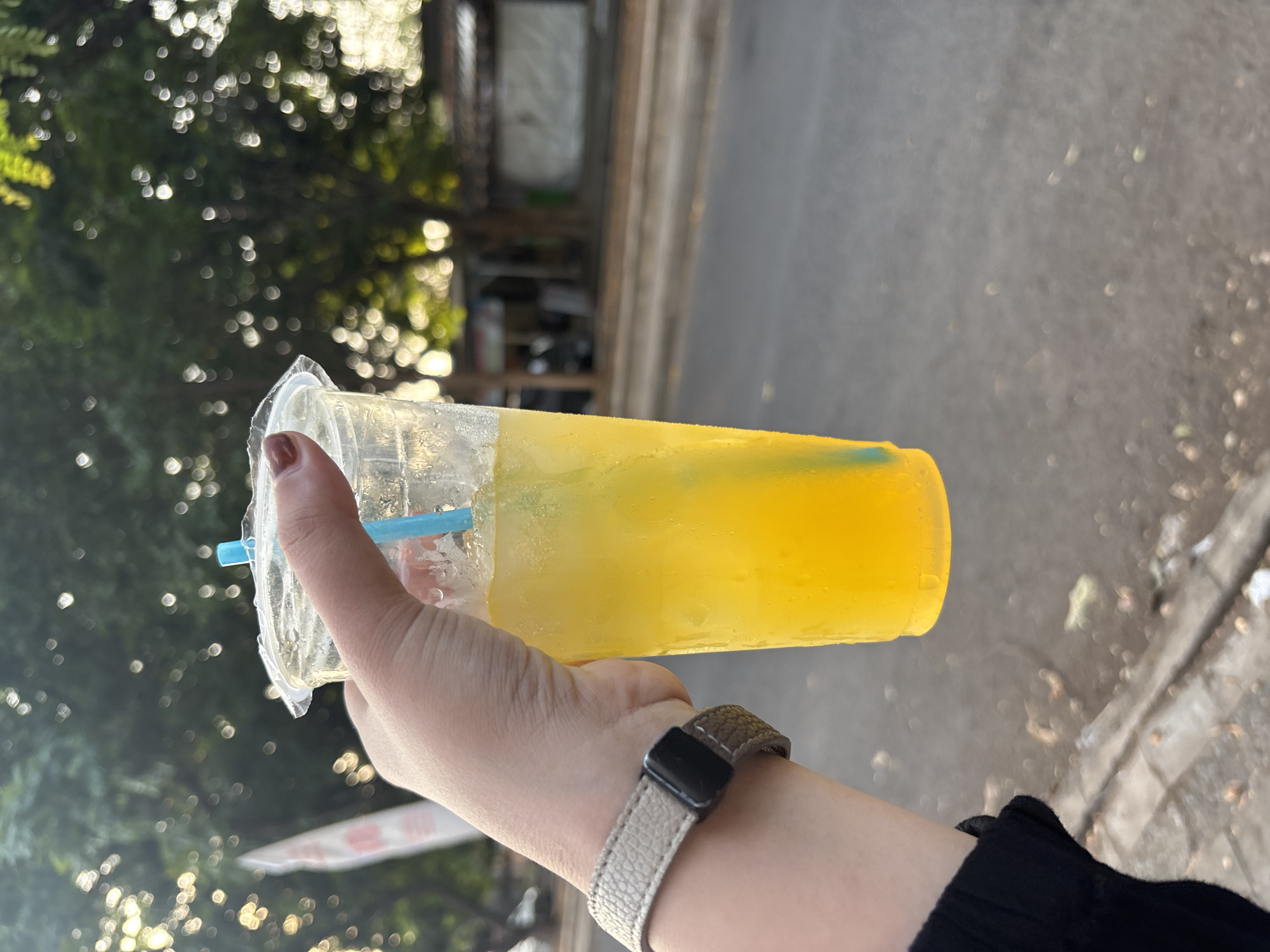 A person's hand, wearing a light gray textured wristband and a black smartwatch, holds a clear plastic cup filled with a bright yellow, ice-filled beverage. A bright blue straw extends from the cup. The background is blurred, showing a street scene with trees and buildings, suggesting an outdoor setting. The overall lighting indicates it is daytime. Given the context of Cirebon, Indonesia, this scene could depict a person enjoying a refreshing drink on a street in the city.