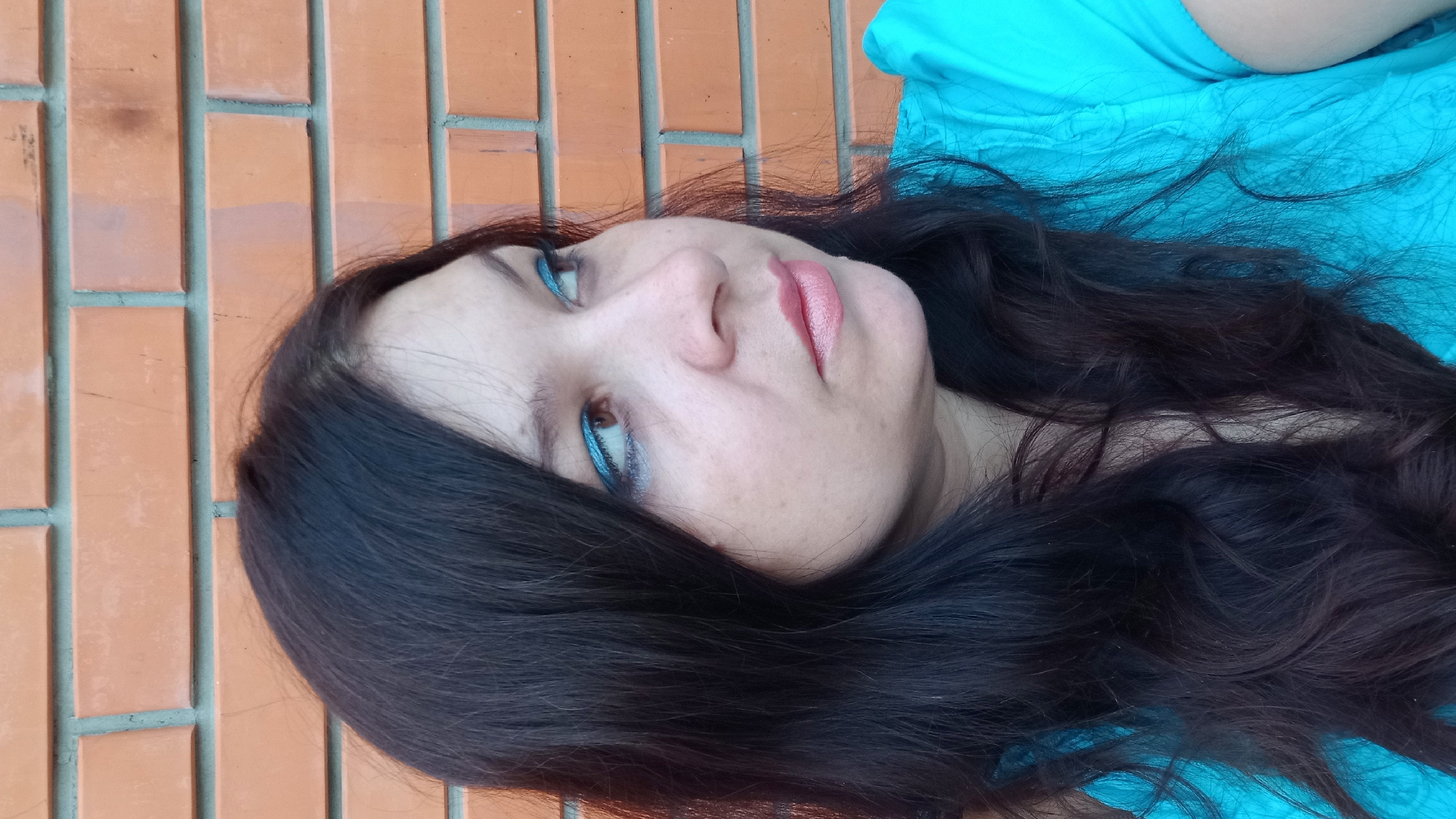 In Kropyvnytskyi, Ukraine, a woman with dark, wavy hair is captured from the chest up, her face turned to the right, looking away from the camera. She is wearing a bright turquoise top with a textured pattern. Her makeup includes shimmering blue eyeshadow and light pink lipstick. She stands in front of a brick wall, the bricks being orange-red and laid in a horizontal pattern. The lighting suggests it is daytime, with natural light illuminating her face.