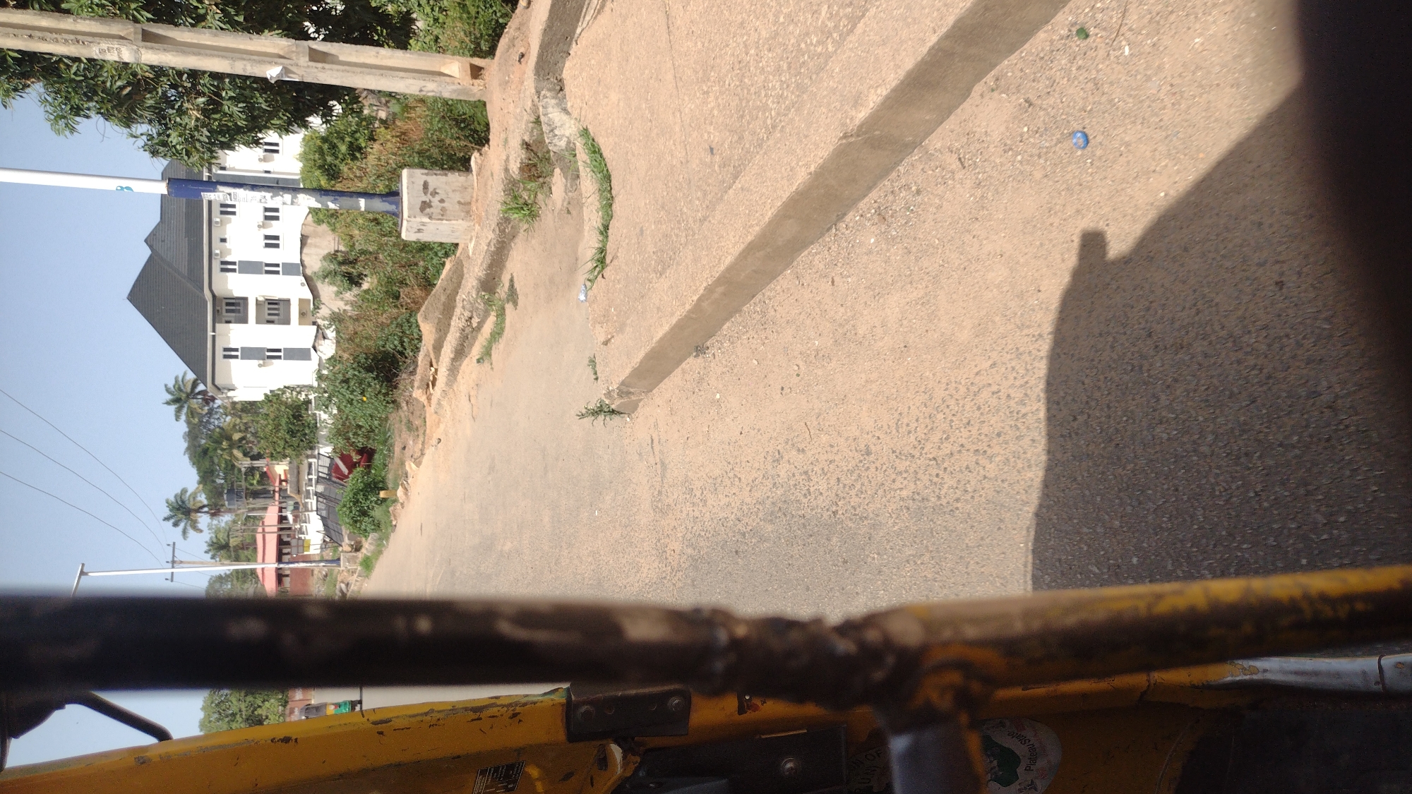 The image captures a street view, likely taken from inside a vehicle moving along the road in Jos, Nigeria. The road is paved and slightly worn, with a concrete curb and a narrow drainage channel running alongside it. Some vegetation grows along the curb. A blue plastic bottle rests on the edge of the road.
In the background, a two-story white building with a dark roof is visible. Other buildings, trees, and utility poles line the street. The sky is clear and blue.
The lower portion of the frame shows parts of the vehicle, including yellow metal bars. The vehicle appears to be a tricycle (Keke Napep).