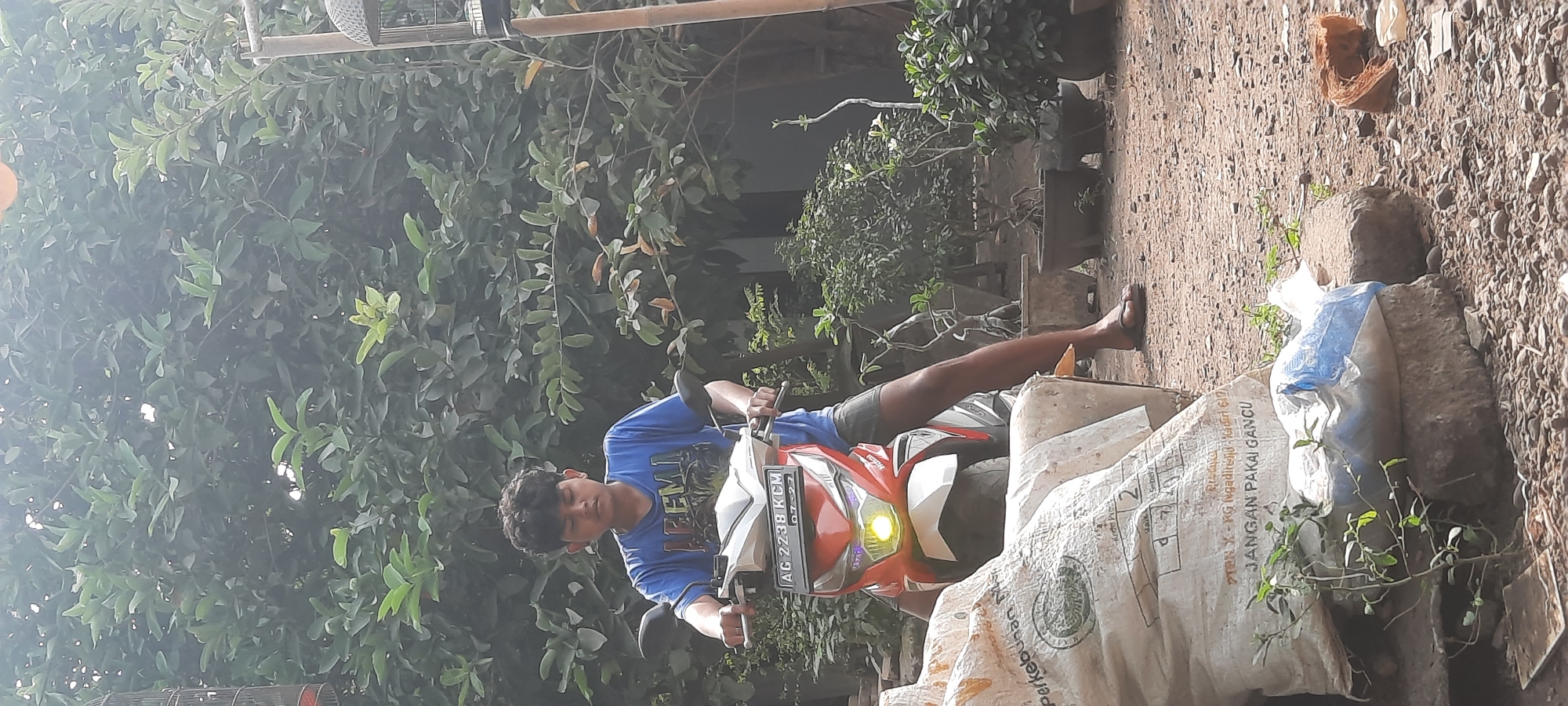 A young person with curly dark hair, wearing a blue t-shirt and dark shorts, is seated on a red and white scooter. The scooter has a license plate that reads "AG 2238 KCM" and the date "07-27". The person is holding the handlebars. The scooter's headlight is on, emitting a yellow glow.

The background consists of lush green foliage, with trees and smaller plants visible. To the right, a birdcage is suspended from a wooden pole. In the foreground, there are large, light-colored sacks, one of which has text that is partially obscured but includes the phrase "JANGAN PAKAI GANCU". There are also rocks, gravel, and a plastic bag containing white material. The ground appears to be dirt and small stones. The scene is outdoors, suggesting a residential area or yard. The overall impression is a casual, everyday moment captured in Selopuro, Indonesia.