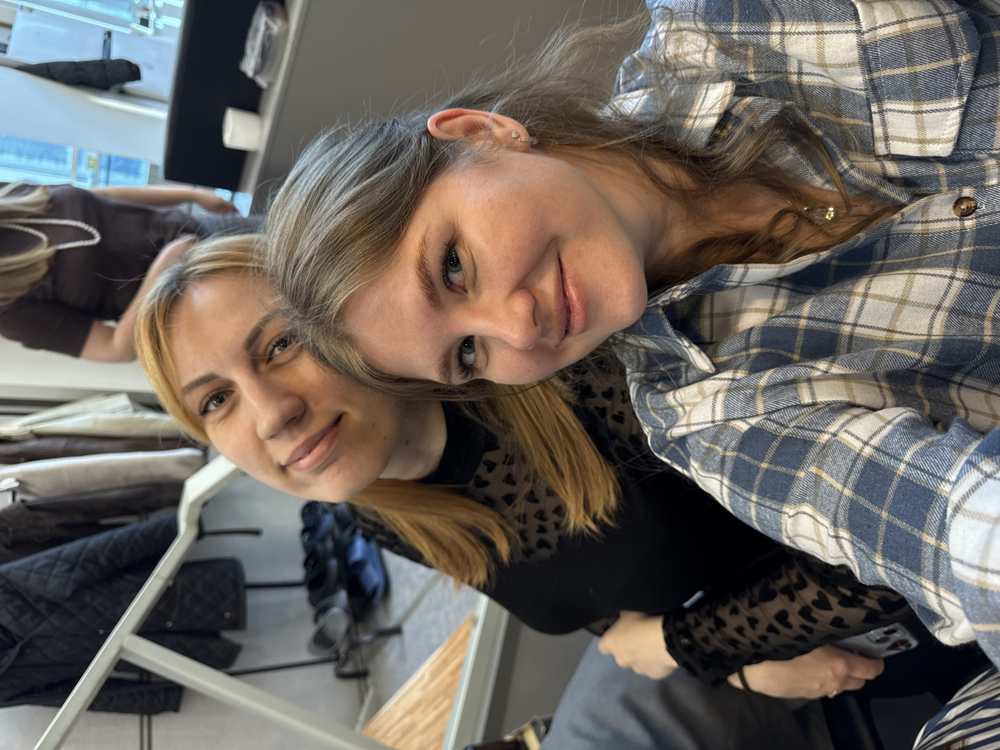 Two women are posing for a selfie in what appears to be a clothing store. The woman on the left has blonde hair and is wearing a black top with a heart pattern on the sleeves. The woman on the right has blonde and brown hair and is wearing a blue and white plaid shirt. In the background, there are clothes racks with various jackets and other garments. The location is inferred to be in Perm, Russia.