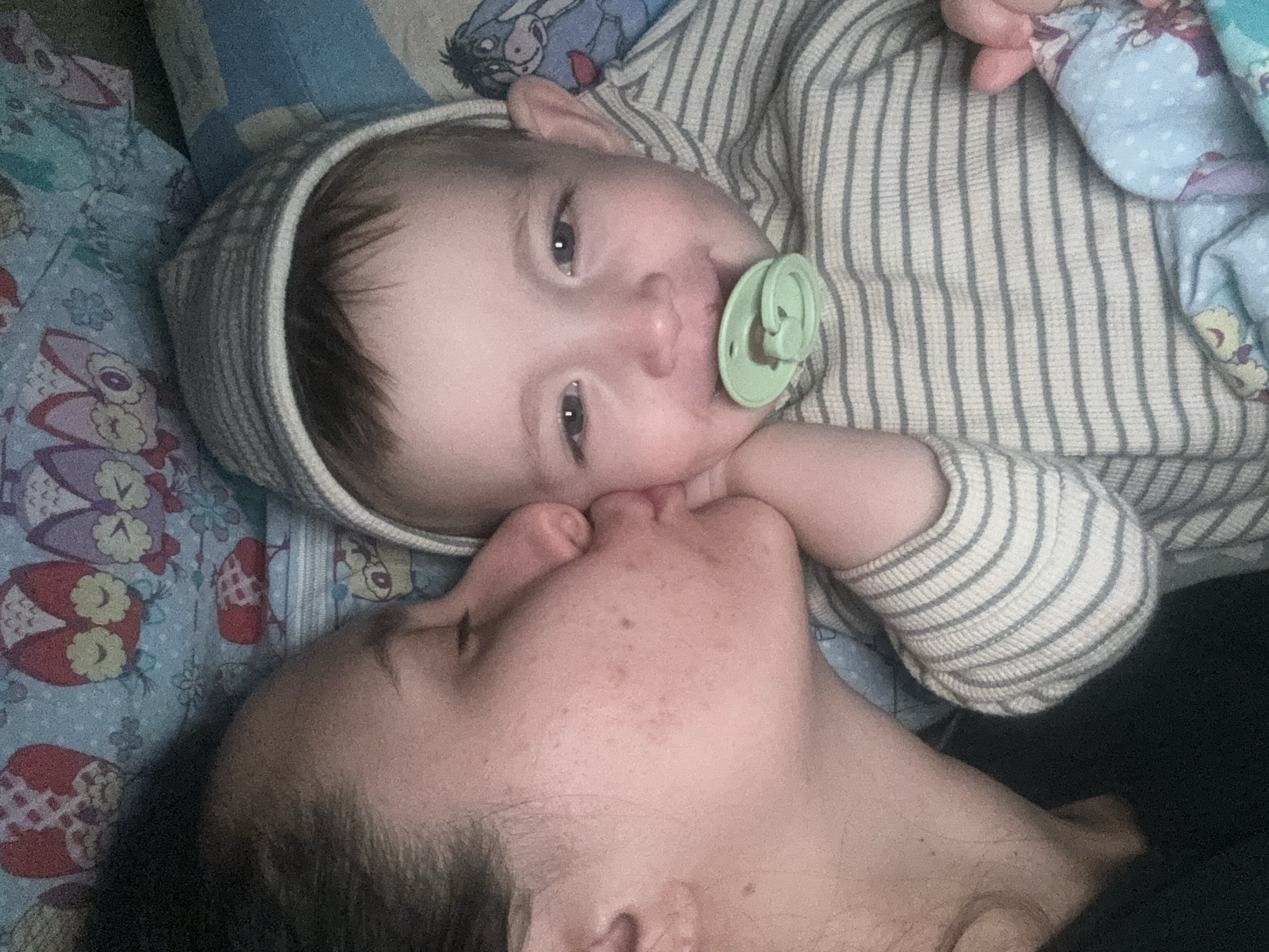 A close-up, medium shot captures a baby and a woman in an intimate moment. The baby, wearing a striped hooded garment and having a green pacifier in its mouth, lies on patterned bedding. The woman's face is positioned near the baby's, their noses touching, indicating a kiss. The background includes patterned fabrics, possibly bedding or blankets. No specific information indicates the location is in Красный Маяк, Russia. The scene focuses on the interaction between the baby and the woman, emphasizing a loving and familial connection.