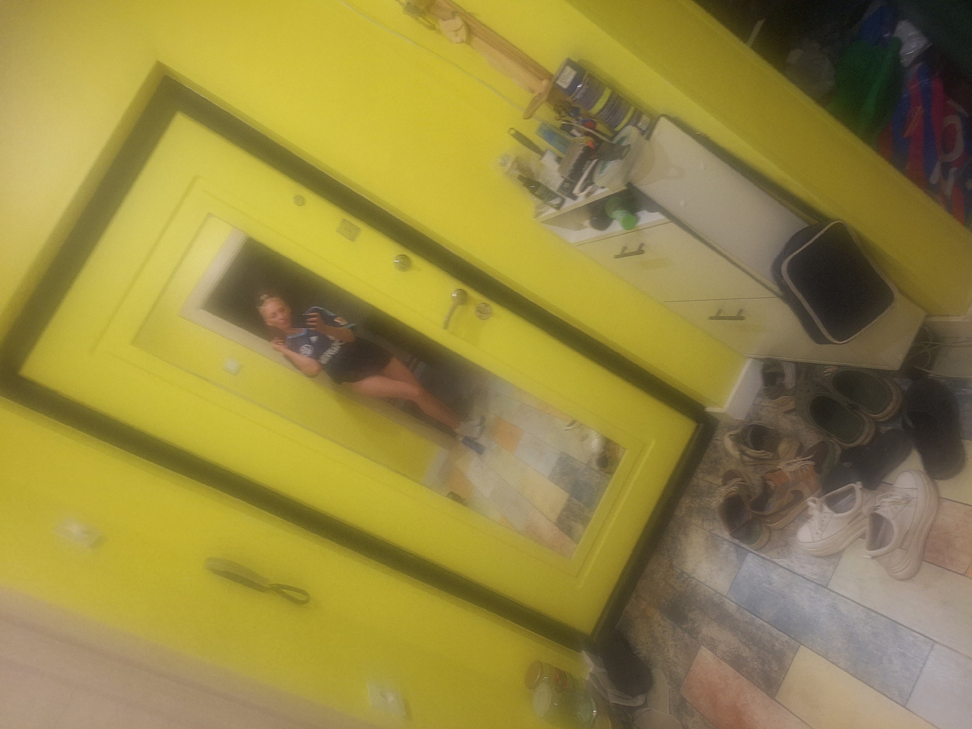 A full-body selfie is taken in a mirror, showing a woman in a black top and dark shorts standing in a brightly lit hallway. The hallway walls are a vivid yellow, and a dark-framed door with a large mirror dominates the background. The mirror reflects the woman, who is holding a smartphone to take the picture. The floor is tiled with a mosaic of colors. To the right of the door is a white cabinet with drawers, on which various items are placed, including bottles and canisters. A wooden object is mounted on the wall above the cabinet. In the foreground, a collection of shoes is scattered on the floor, including sneakers and boots. The overall scene depicts a casual indoor environment, possibly a home or an apartment entrance. The image does not provide any specific visual cues to pinpoint its location in Kaluga, Russia.