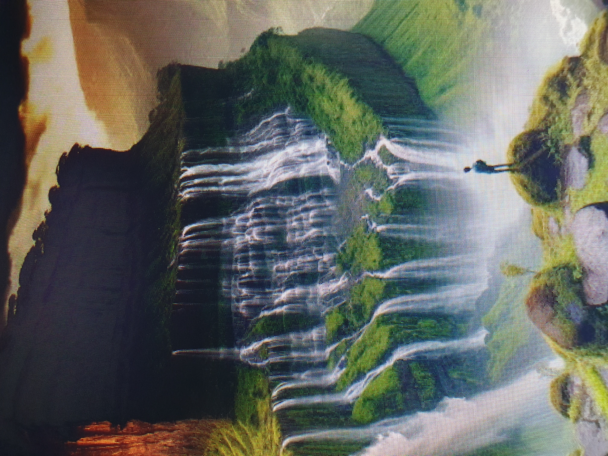 The image depicts a vast, multi-tiered waterfall cascading down a sheer cliff face. The cliff is covered in lush green vegetation, creating a vibrant contrast with the white water. The scale of the waterfall is immense, dwarfing a lone figure standing on a rocky outcrop in the foreground. The figure, seen from behind, appears to be a person wearing a blue shirt and dark pants, gazing out at the majestic landscape. The sky above is partly cloudy, with warm, golden light illuminating the scene, suggesting either sunrise or sunset. The environment is wild and natural, characterized by dramatic geological formations and abundant greenery. There is no indication that this scene is located in Tashkent, Uzbekistan; it appears to be a natural landscape.