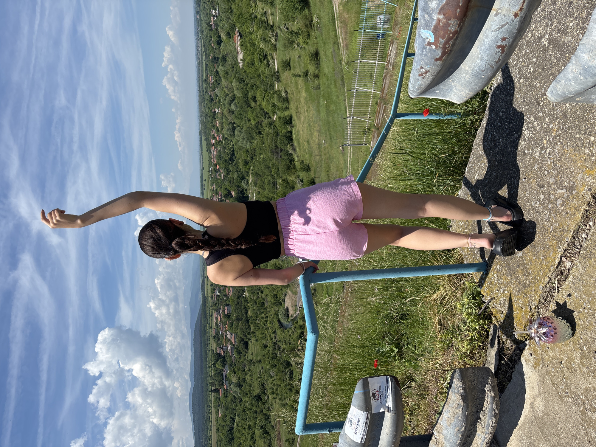 This image captures a young woman standing at a scenic overlook, gazing upon a vast landscape. The photograph is oriented vertically, showing the woman in a black tank top and pink shorts, with her dark hair styled in a braid. Her left arm is extended upwards, while her right hand rests on a blue metal railing, seemingly posing or stretching while enjoying the view.
The setting is a high vantage point, with a concrete path in the foreground and patches of green grass. A vibrant blue sky with scattered white clouds dominates the upper half of the image. Below, a sprawling panorama unfolds, featuring dense green forests, cultivated fields, and distant villages with small houses. Mountains are faintly visible on the horizon, adding depth to the scene. The bright sunlight indicates a daytime shot, likely mid-day, as evidenced by the distinct shadow of the woman cast on the concrete ground.
Notable objects include the sturdy blue metal railing, which appears to be part of a protective barrier. On the concrete, near the railing, a small decorative item resembling a faceted pineapple or strawberry ornament is visible. A white plastic bag or box, attached to the railing, displays the Cyrillic text "АРУС СПОРТ" (ARUS SPORT). The overall atmosphere suggests a peaceful moment appreciating the natural beauty of the Bulgarian landscape, specifically in Ognyanovo.